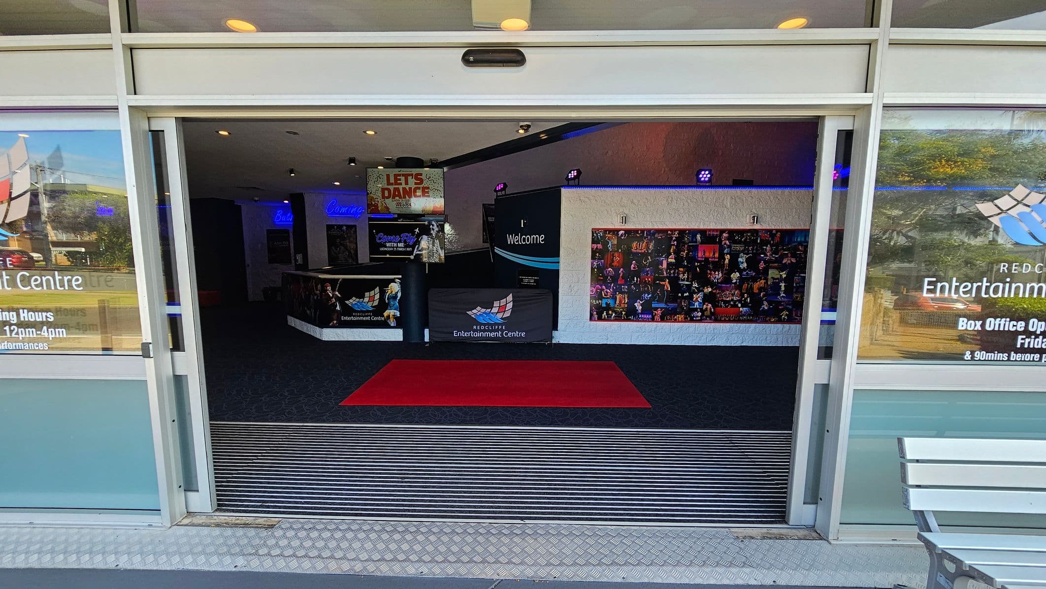 Entrance to a building with glass doors open, revealing an interior with a red carpet on a dark floor. The left and right sides have posters and signage, including "Redcliffe Entertainment Centre" and box office hours. Inside, a "Welcome" sign and a collage of images are visible on the walls. The ceiling has recessed lighting, and a bench is partially visible on the right outside.