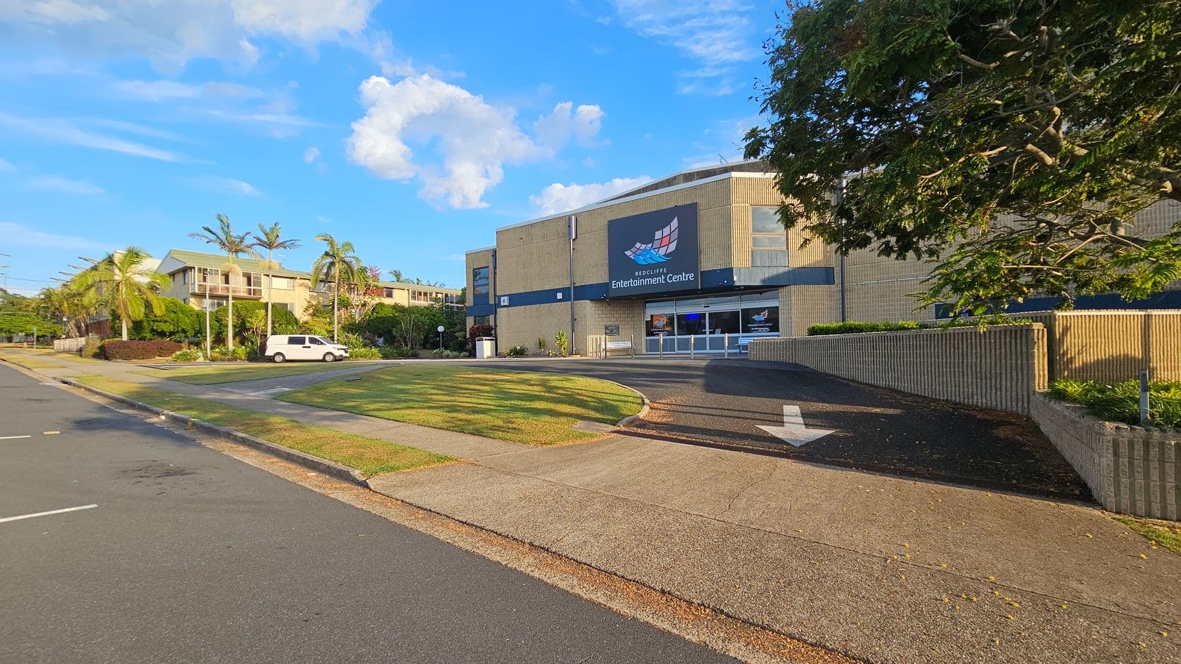 A street view with a large building on the right labelled "Redcliffe Entertainment Centre". The building is beige with a dark blue sign. A tree with green leaves partially shades the building. On the left, a white van is parked near a row of palm trees and residential buildings. The foreground features a paved footpath and a grassy area, with a road running alongside. The sky is clear with a few clouds.