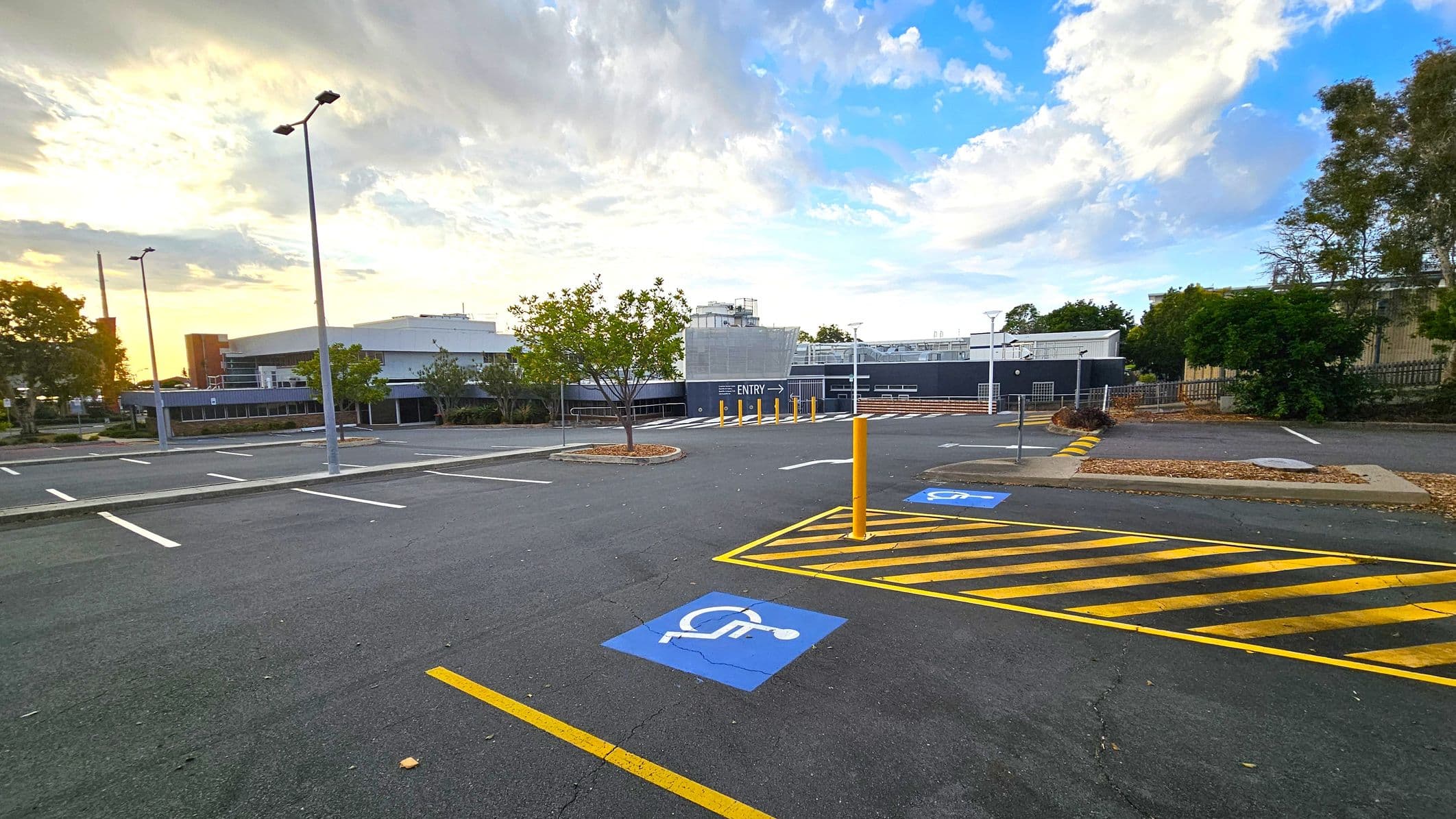 An empty car park with marked parking spaces, including a blue disabled parking sign on the ground. Yellow-striped lines are visible near the foreground. Several tall streetlights are scattered throughout the area. In the background, there is a large building with a sign indicating "ENTRY" and some trees and shrubs around the perimeter. The sky is partly cloudy with a mix of blue and light hues.