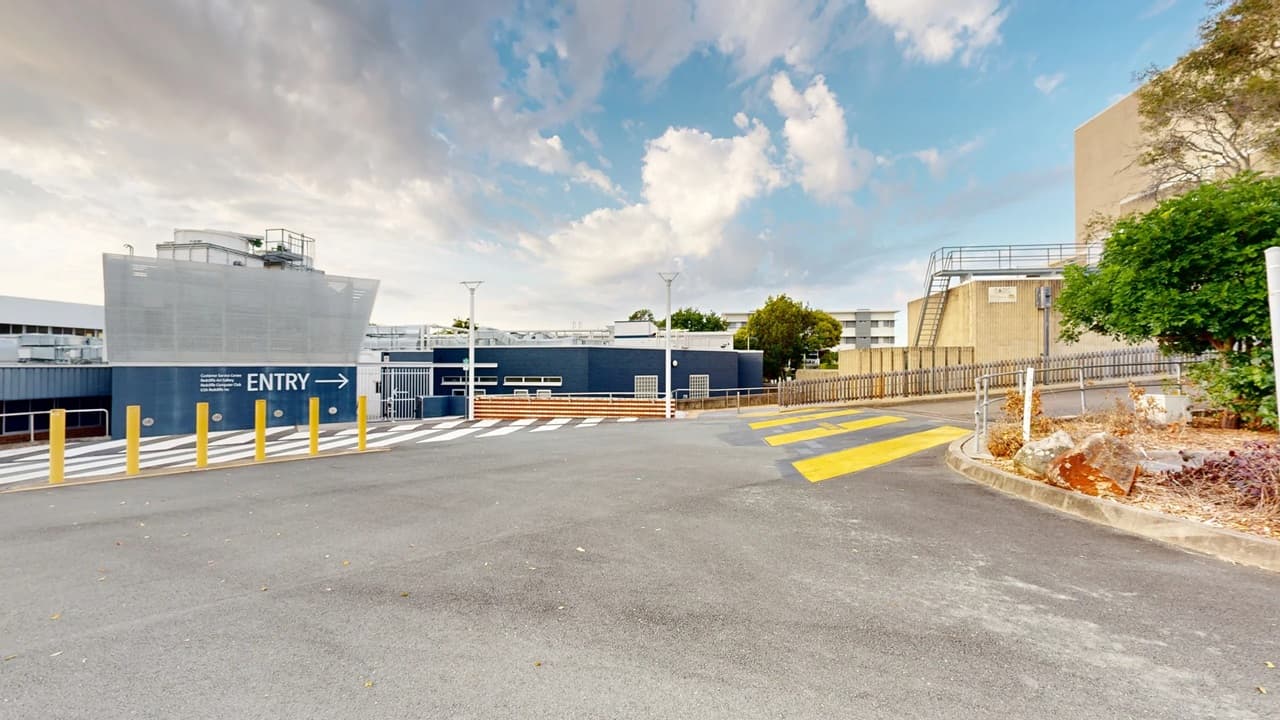 A paved area with a marked pedestrian crossing in yellow and white lines. On the left, a blue building with "ENTRY" and an arrow is visible, along with several yellow bollards. In the background, there are trees and additional buildings with metal railings. On the right, a small landscaped area with rocks and shrubs is present. The sky is partly cloudy.