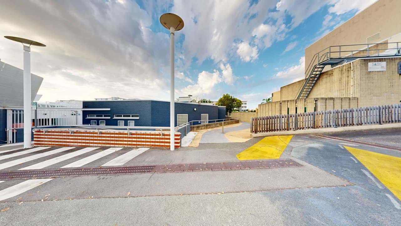 A paved area with a zebra crossing in the foreground. On the left, there is a blue building with white-framed windows and a red and white striped barrier. Two tall street lamps are visible. On the right, a beige building with a metal staircase and a wooden fence. The background features a tree and some distant buildings. The ground has yellow painted markings, and the sky is partly cloudy.