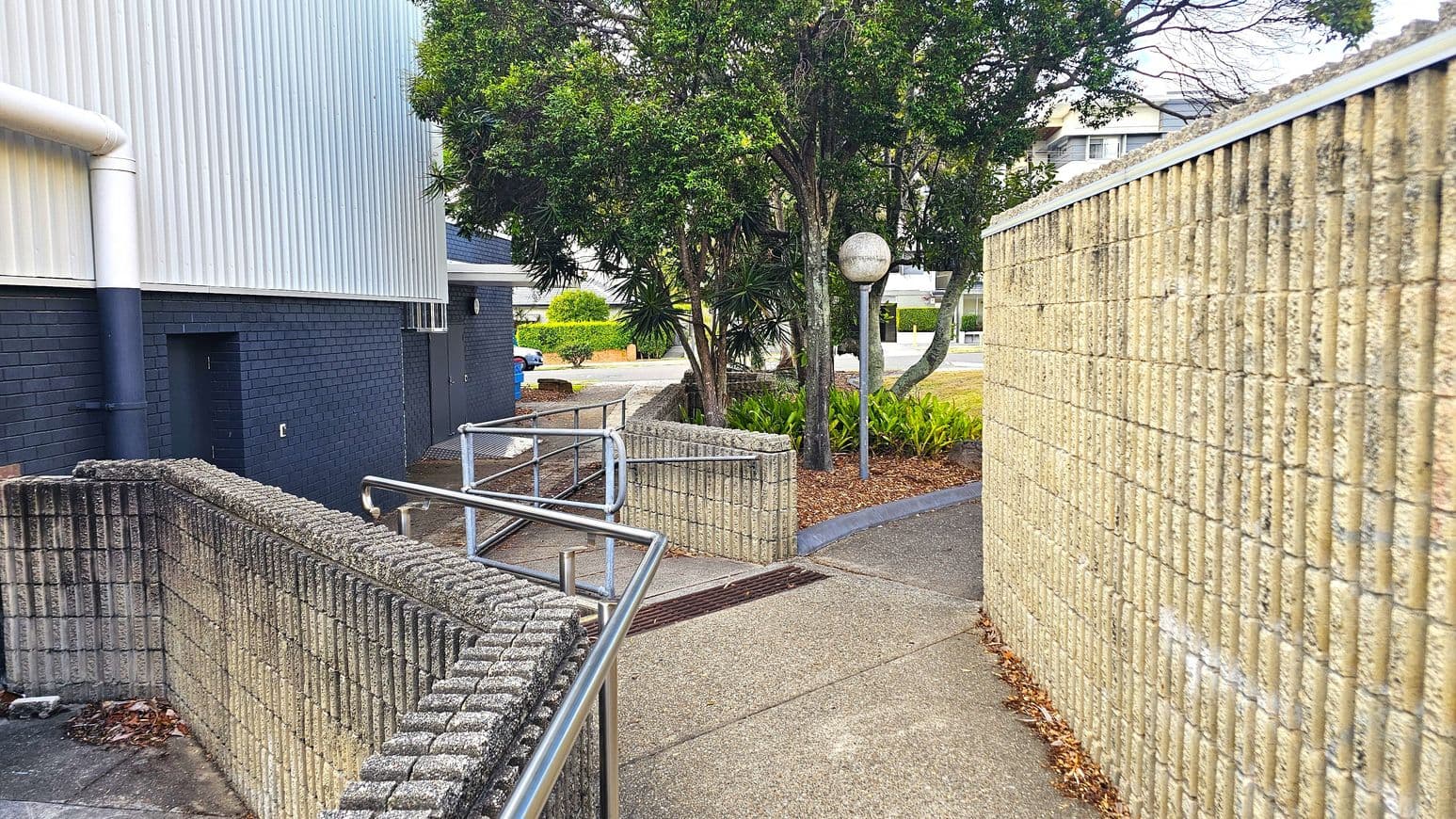 A narrow pathway with a textured concrete wall on the right and a metal railing on the left. The ground is paved with concrete. In the middle, there are several trees and a spherical streetlamp. The background shows a building with a corrugated metal exterior and a dark brick wall. Some greenery and a parked car are visible further back.