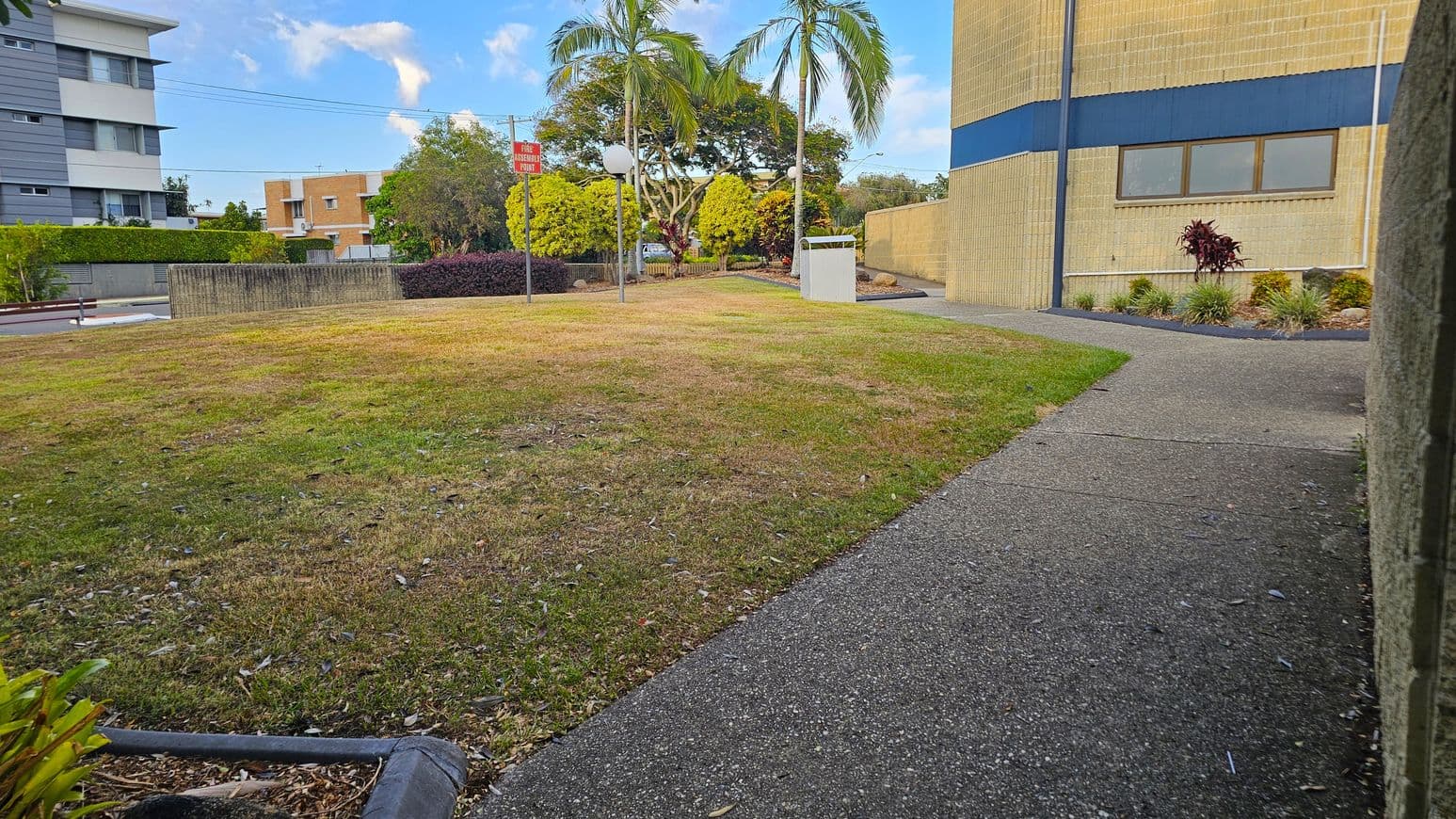 A grassy area with patches of brown is in the foreground, bordered by a concrete path on the right. In the background, there are several trees, including palm trees, and a few buildings. A signpost and a white structure are visible near the centre. The building on the right has a beige and blue exterior with windows, while another building on the left is grey and white. Bushes and small plants are along the path.