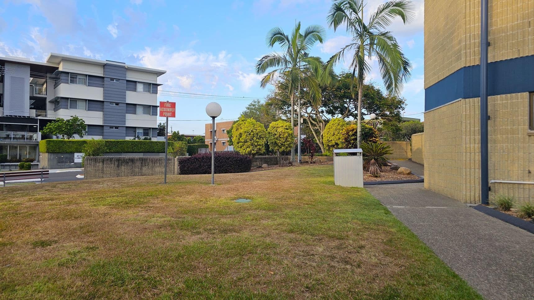 A grassy area with patches of brown is in the foreground. On the left, a modern building with grey and white horizontal panels is visible. A "Fire Assembly Point" sign and a spherical streetlamp are near the centre. On the right, a beige building with a blue stripe is partially visible. Several palm trees and bushes are in the background, along with a small pathway and a bin. A low brick wall separates the grassy area from the road.