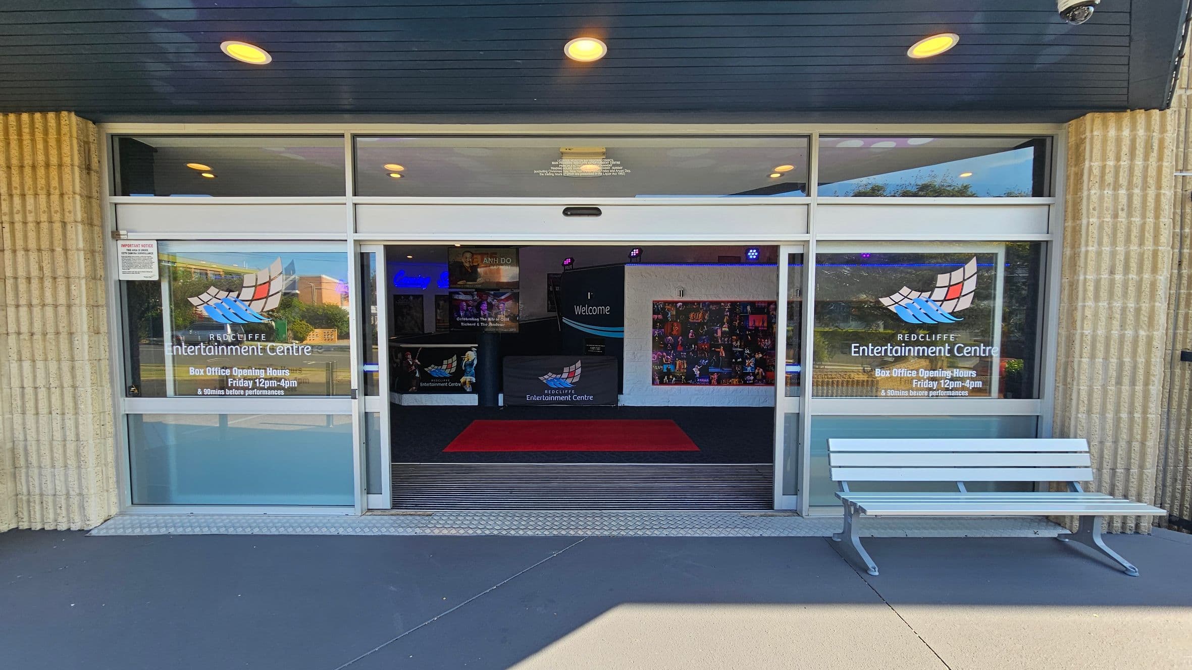Entrance to a building with large glass doors and windows. The windows display the logo and name "Redcliffe Entertainment Centre" along with box office hours. A white bench is positioned on the right side of the entrance. Inside, a red carpet leads to a welcome sign and colourful posters on the walls. The ceiling is dark with recessed lighting, and the floor outside is grey.