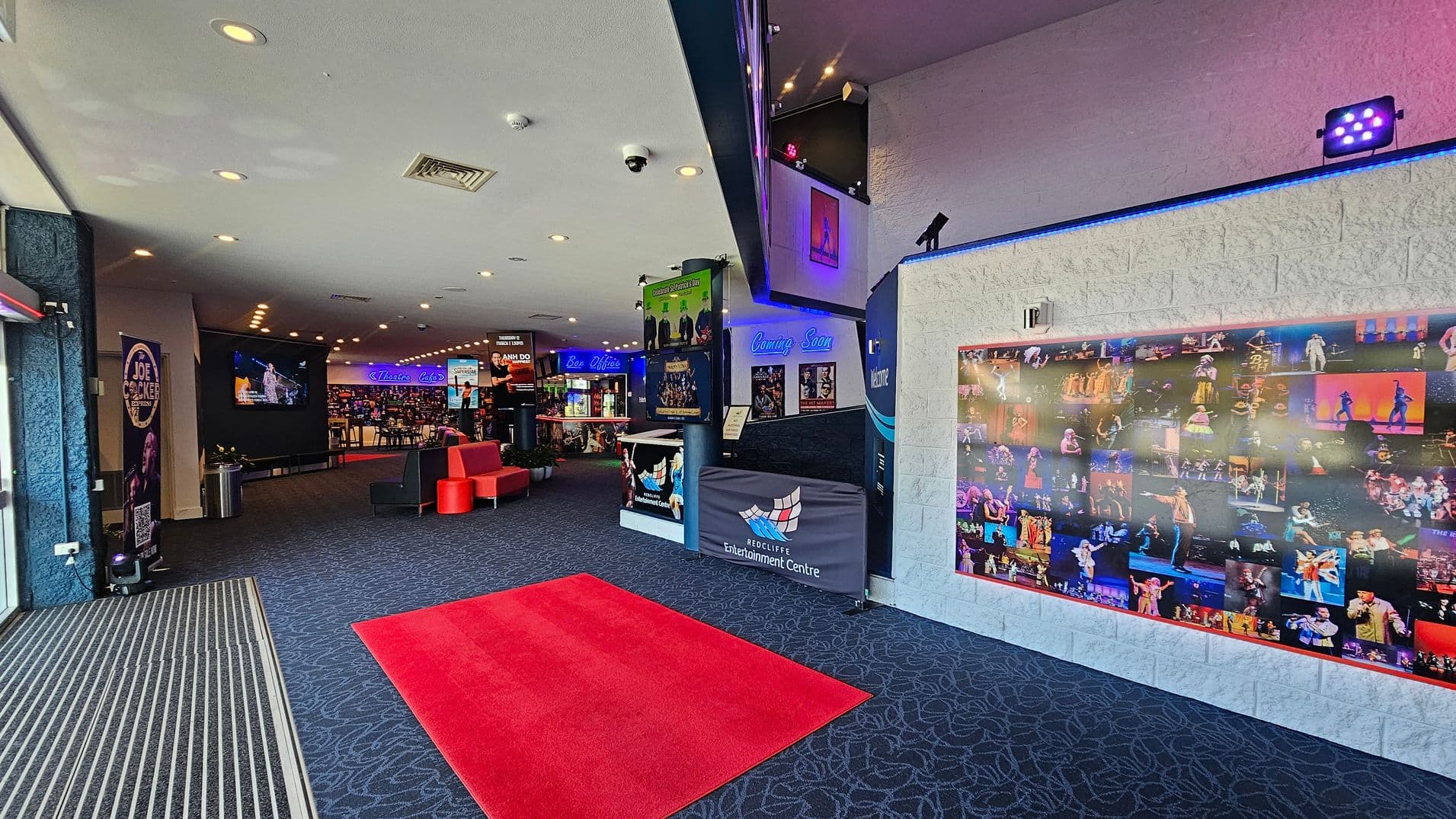 Entrance area with a patterned dark carpet and a red mat leading inside. On the left, a large screen displays a performance, with a poster and a bin nearby. In the centre, a seating area with red and black chairs is visible. The right wall features a collage of performance photos. Above, a sign reads "Coming Soon" with various posters. The ceiling has recessed lighting and a camera.
