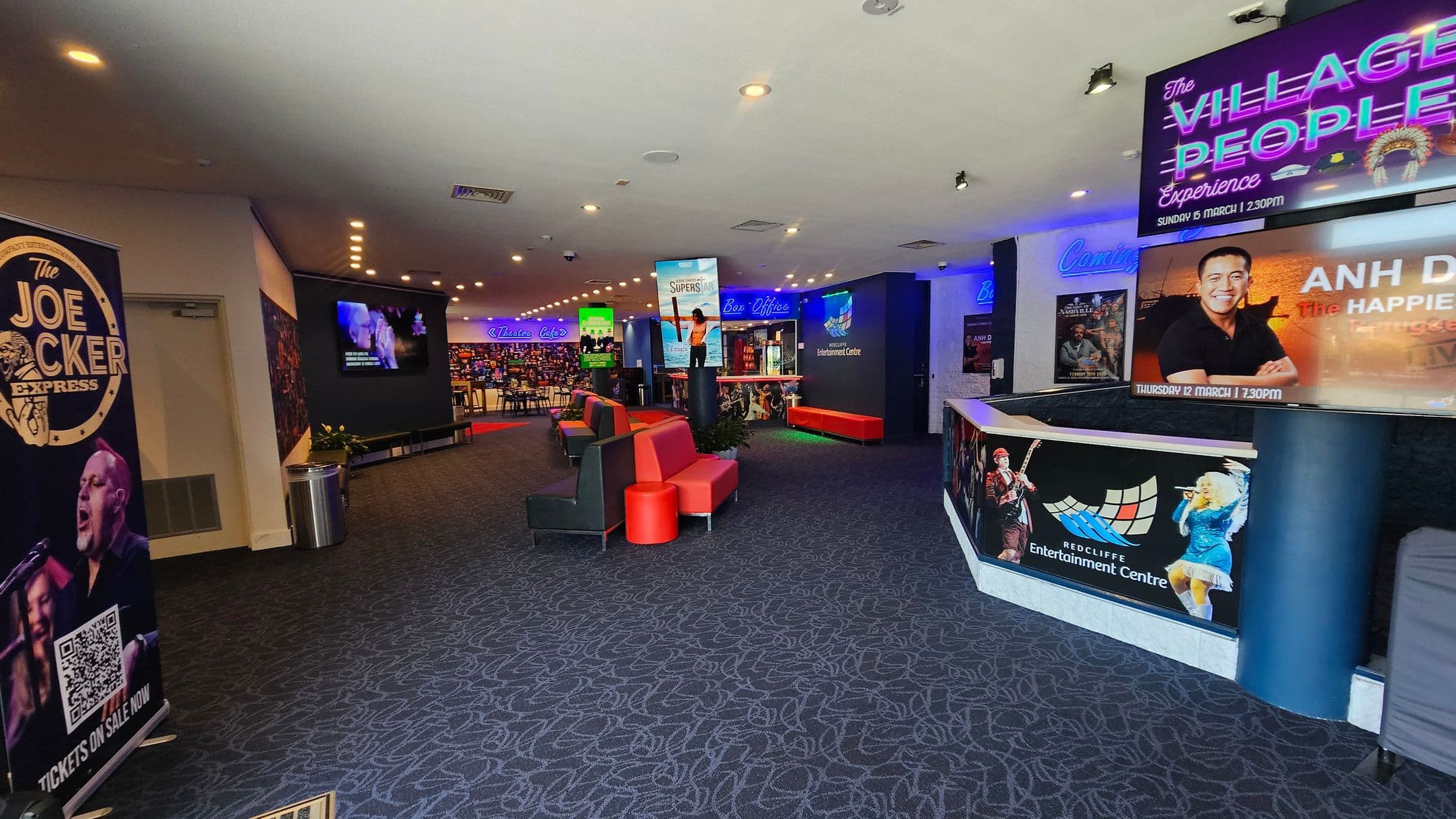 A spacious lobby with a patterned carpet floor. On the left, a large poster for "The Joe Cocker Express" is displayed. In the centre, red and black seating is arranged, with a digital screen showing promotional content. The right side features a counter with posters for upcoming shows, including "The Village People Experience" and "Anh Do: The Happiest." Neon signs for "Theatre" and "Box Office" are visible in the background, along with a wall of colourful lights. The ceiling has recessed lighting.