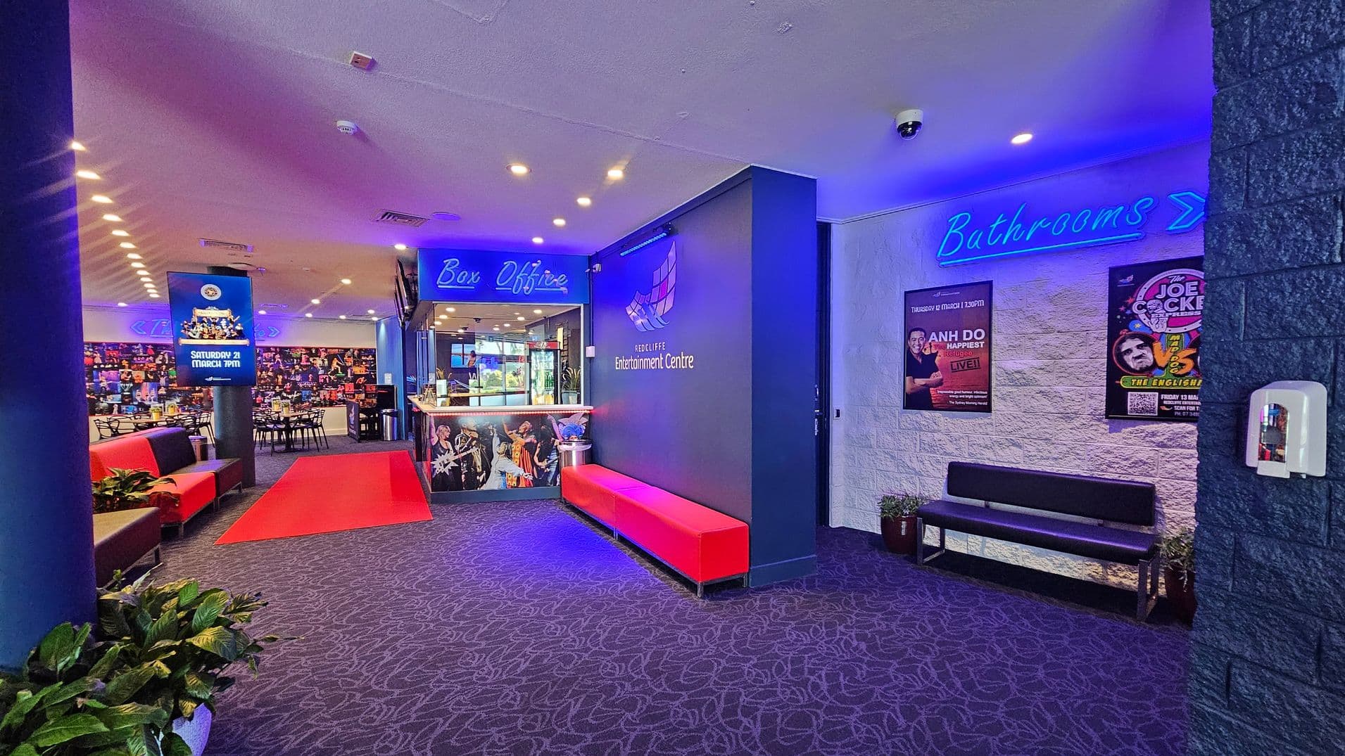 A vibrant entertainment centre lobby with a patterned carpet. On the left, red and black seating lines the wall, with a digital display advertising an event. In the centre, a red carpet leads to the box office, which is illuminated with blue signage. The right wall features posters and a blue neon "Bathrooms" sign above a black bench. Potted plants are placed around the area, and a hand sanitiser dispenser is mounted on the wall. The ceiling has recessed lighting.