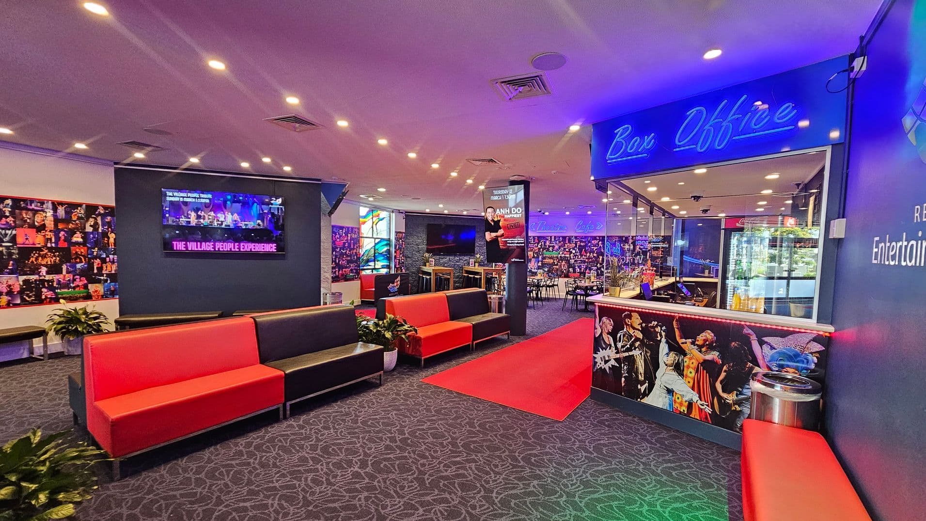 A vibrant lobby area with a patterned carpet and a ceiling dotted with recessed lights. On the left, a wall features a collage of performance images and a screen displaying "The Village People Experience." Red and black cushioned benches are arranged along the wall. On the right, a box office with a neon sign and a counter decorated with performer images. In the background, a café area with tables and chairs is visible, along with a red carpet leading towards it.