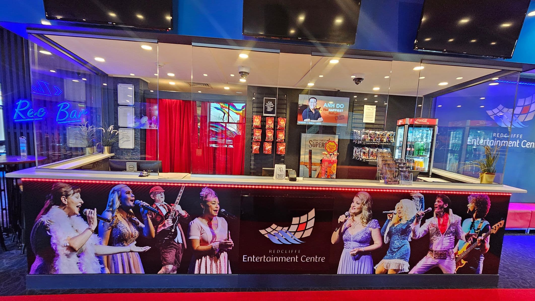 A ticket counter at the Redcliffe Entertainment Centre with a glass partition. The counter features images of performers and the centre's logo. Behind the counter, there are red curtains, a display of snacks, and a refrigerator with drinks. A digital screen shows an advertisement. To the left, a neon sign reads "Rec Bar" with seating visible. The floor is carpeted in dark tones, and the ceiling has recessed lighting.
