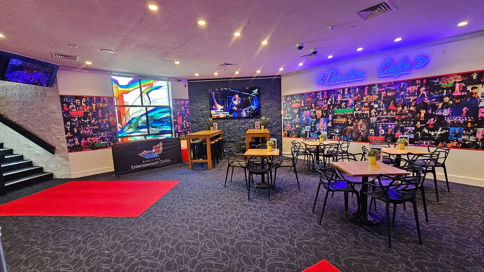 A café area with a patterned carpet and several wooden tables surrounded by black chairs. On the right, a wall is covered with a collage of performance photos and a neon sign reading "Theatre Cafe." In the centre, a large screen displays a performance. To the left, a stained glass window adds colour, and a staircase leads upwards. A red carpet is placed on the floor near the entrance, and a counter with "Redcliffe Entertainment Centre" signage is visible. Ceiling lights illuminate the space.