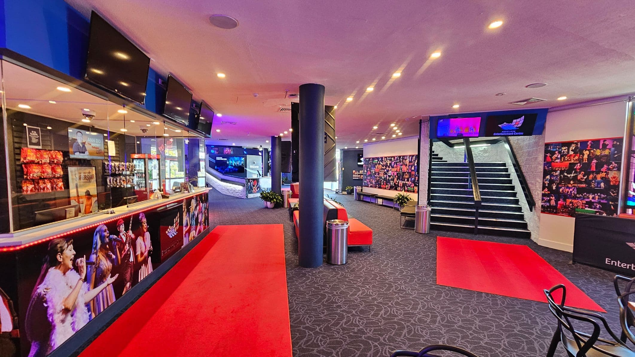 A spacious lobby with a red carpet running through the centre. On the left, there's a counter with a glass display, featuring snacks and promotional posters. Above the counter, several screens are mounted. In the middle, a black column stands between red benches and potted plants. On the right, a staircase leads upwards, flanked by photo collages on the walls. The ceiling is dotted with recessed lights, and the floor is covered with a patterned carpet.