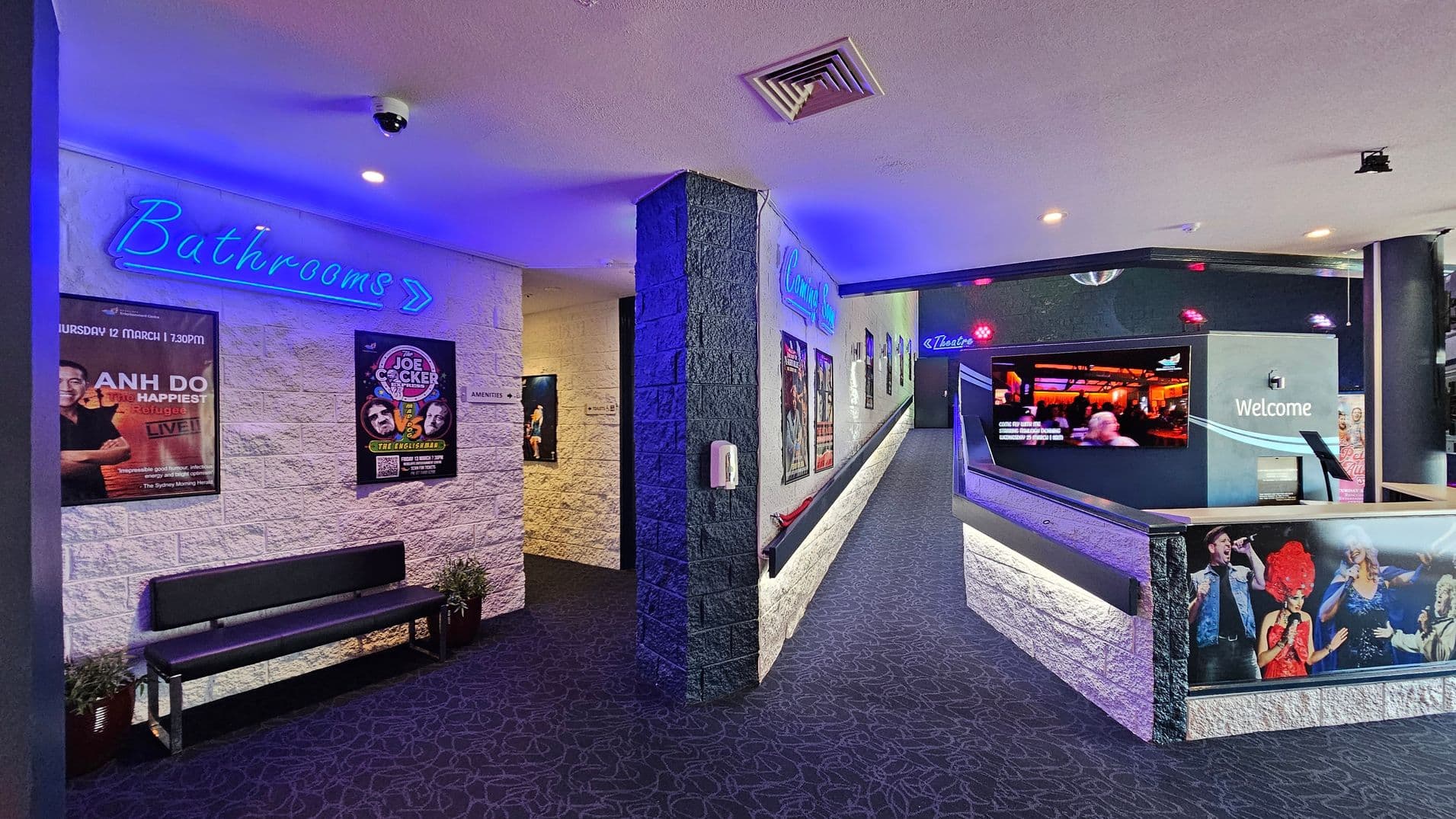 A corridor with a dark patterned carpet and white brick walls. On the left, a blue neon sign reads "Bathrooms" with an arrow pointing right, above a bench and posters. The right side features a ramp leading to a theatre, with a "Welcome" sign and a large screen displaying images. Posters and a neon "Theatre" sign are visible along the wall. The ceiling is light-coloured with a security camera and vents.