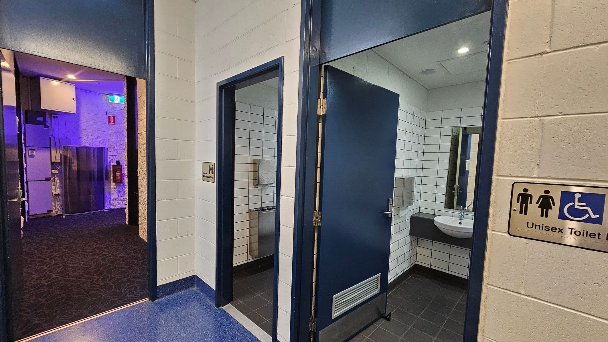 A corridor with two open doors leading to unisex toilets. The left door reveals a toilet with a paper dispenser and a stainless steel fixture. The right door shows a sink with a mirror above it. Both rooms have white tiled walls. The floor is dark, and the ceiling has recessed lighting. A sign on the right wall indicates the toilets are unisex and accessible. The corridor outside has a dark carpet and a blue-lit area with visible electrical panels.