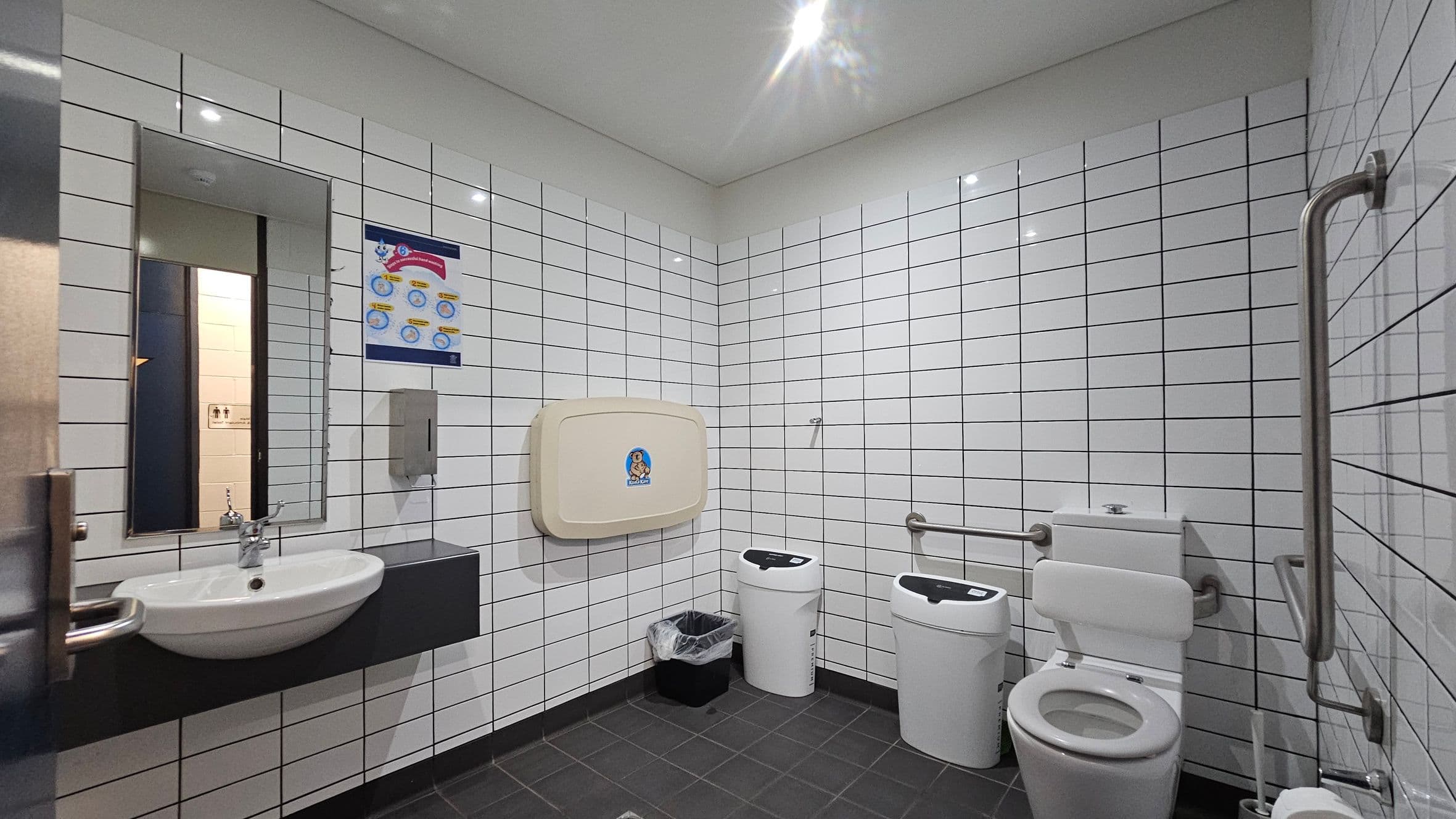 A public restroom with white tiled walls and a dark tiled floor. On the left, a sink with a mirror above it and a soap dispenser nearby. In the centre, a baby changing station is mounted on the wall, with a poster above it. Below, a small bin with a liner. On the right, a toilet with support rails and two sanitary bins. The ceiling is plain with recessed lighting.
