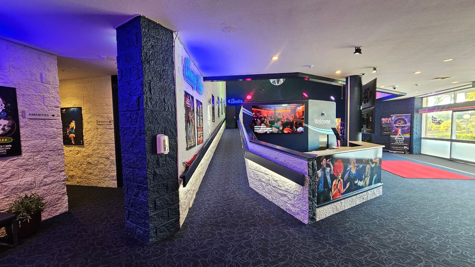 A theatre lobby with a patterned carpeted floor. On the left, a corridor leads to amenities and toilets, with posters on the walls. In the centre, a ramp with more posters leads to a theatre entrance. On the right, a reception desk with a "Welcome" sign and images of performers. The ceiling has recessed lighting, and large windows on the right let in natural light. A red carpet is laid out near the windows.