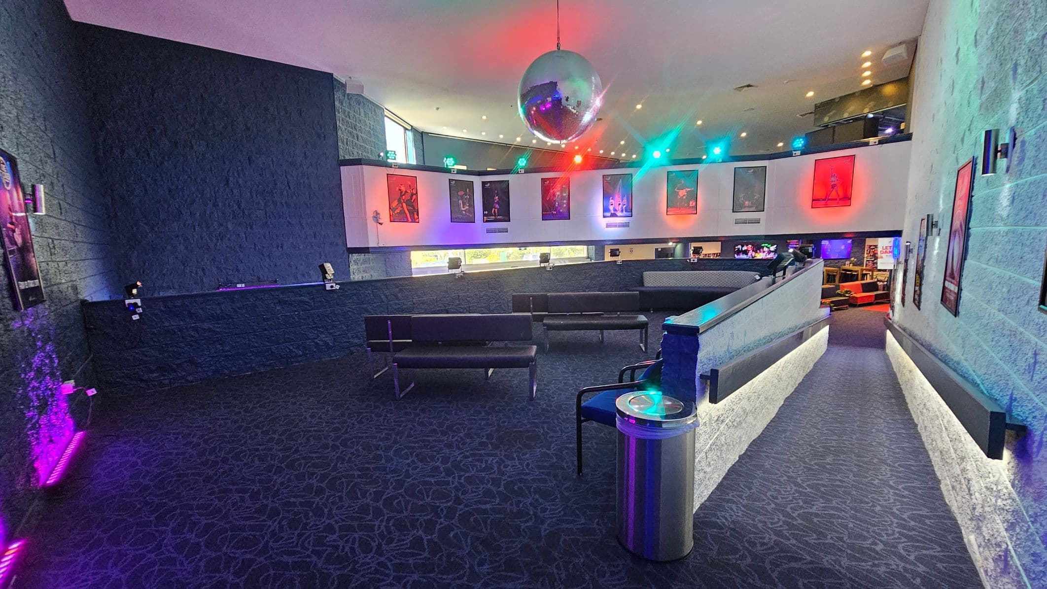 A spacious interior with a dark carpeted floor featuring a swirling pattern. On the left, a textured wall displays several posters, illuminated by purple lighting. In the centre, a large disco ball hangs from the ceiling, casting colourful reflections. Below, black benches are arranged in a seating area. The right side has a sloped walkway with a metal railing and more posters on the wall. The upper level displays additional posters with vibrant lighting effects.