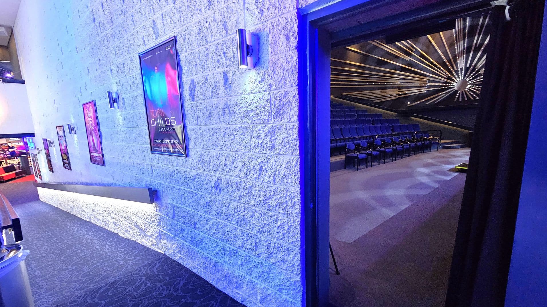 A hallway with a textured white wall on the left, adorned with several posters and modern wall lights. The floor is carpeted with a patterned design. On the right, an open doorway leads into a theatre with rows of blue seats and a striking ceiling design featuring radiating lines and a central circular element. The theatre is dimly lit, contrasting with the brighter hallway.
