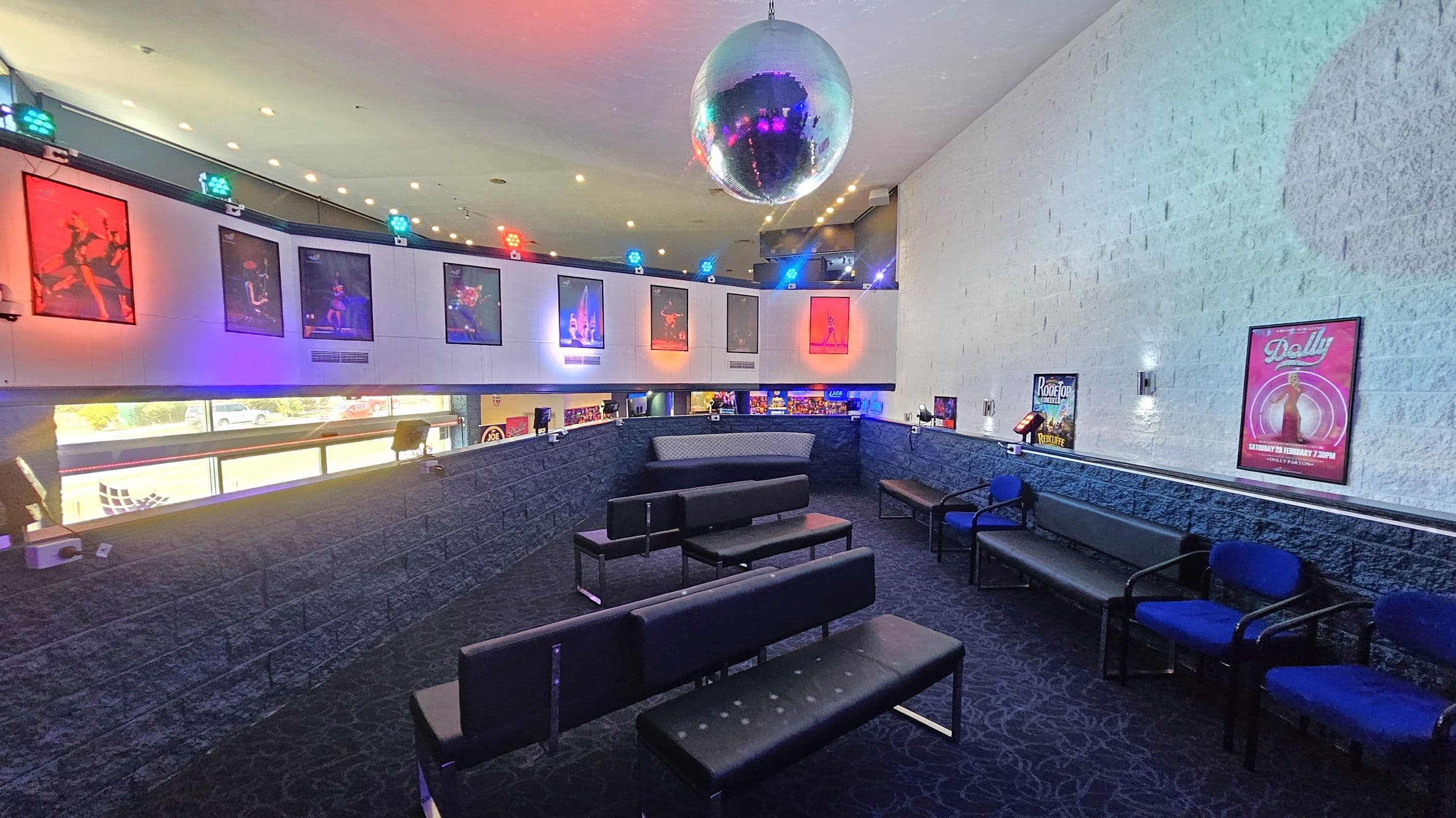 A room with a dark carpeted floor and a white ceiling featuring a large disco ball. On the left, there are colourful posters and lights on the upper wall. The seating area consists of black benches and blue chairs arranged in rows. The right wall is adorned with posters, including one for "Dolly." The lower part of the walls is made of dark stone, and there are windows providing a view of the outside.