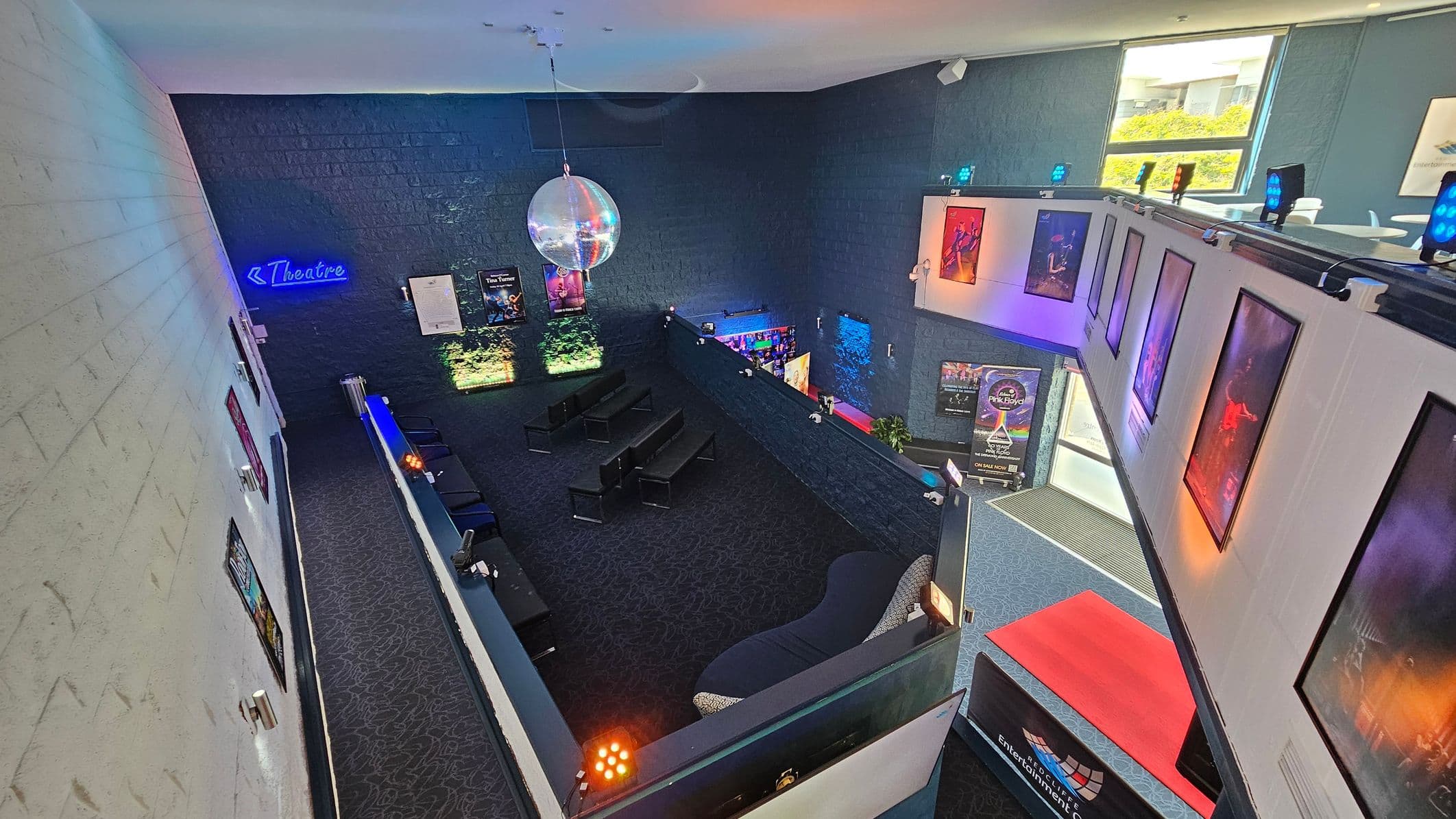 A theatre lobby with a dark carpeted floor and high ceiling. On the left, a white brick wall displays several posters. In the centre, a seating area with black chairs and a large disco ball hanging above. The right side features a balcony with more posters and blue spotlights. A neon sign reading "Theatre" is on the back wall. Bright natural light enters through windows on the upper right, illuminating the space.