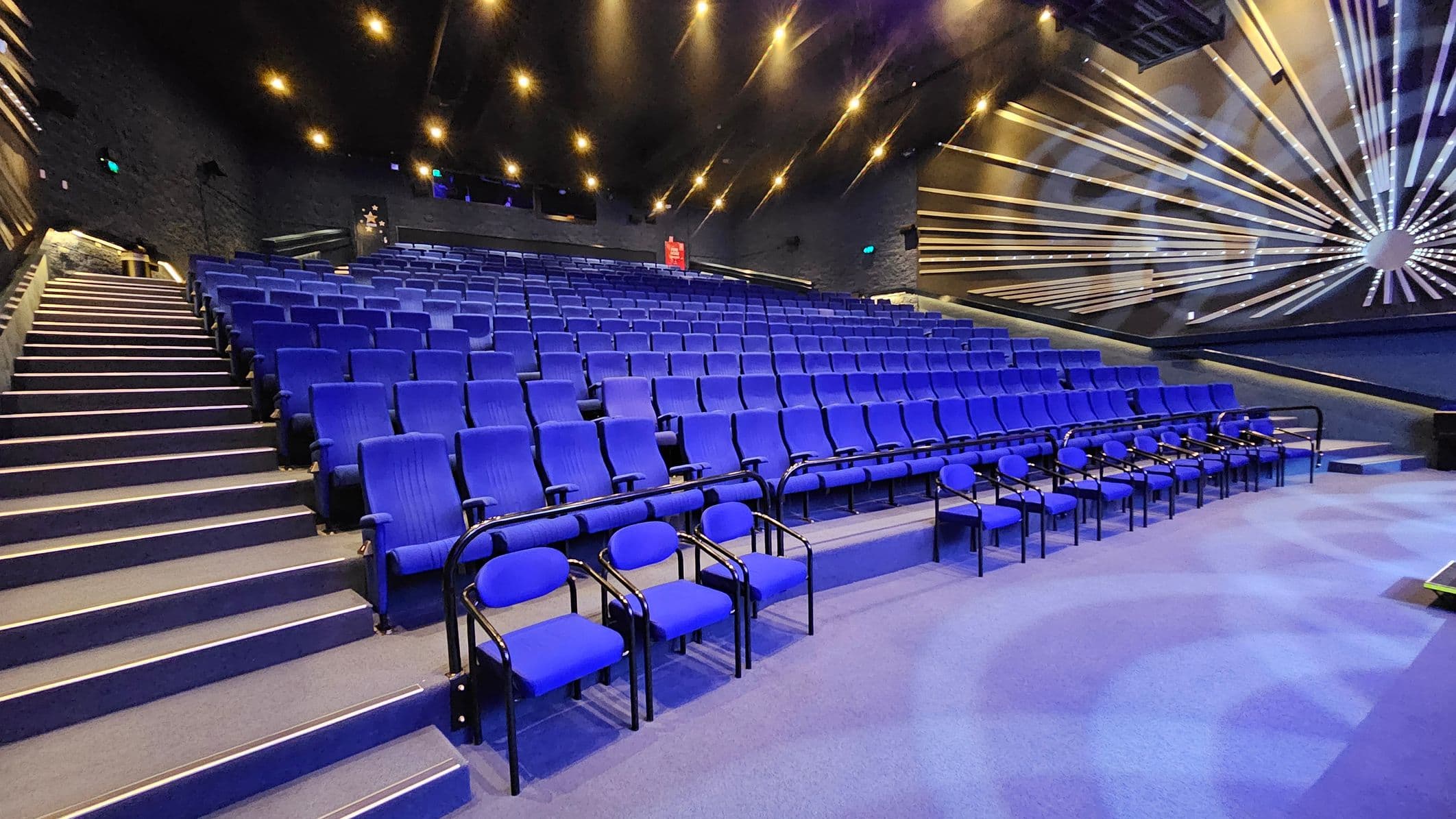 A theatre auditorium with rows of blue seats arranged in tiers. On the left, a staircase with handrails leads up alongside the seating. The right side features a wall with a decorative pattern of radiating lines. The ceiling is dark with numerous small lights, creating a starry effect. The floor is carpeted in blue, matching the seats.