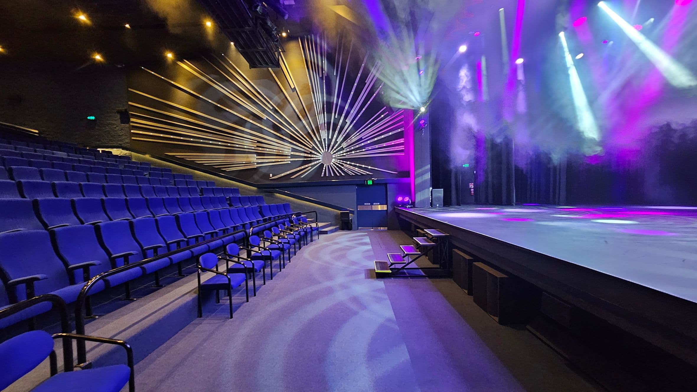 A theatre interior with rows of blue seats on the left, leading up to a stage on the right. The stage is illuminated with colourful lights, casting beams and smoke effects. The back wall features a decorative design with radiating lines. The floor is carpeted, and there are small steps leading up to the stage. The ceiling has spotlights and lighting equipment.