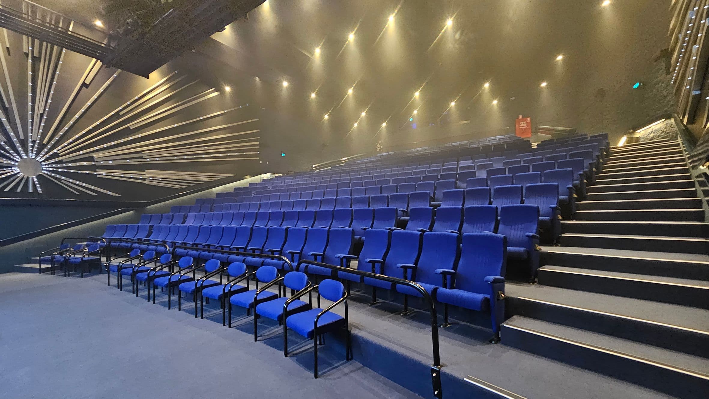 A theatre auditorium with rows of blue seats arranged in a tiered fashion. On the left, there is a striking wall design featuring a radial pattern of metallic lines emanating from a central point. The right side shows a staircase with illuminated steps leading up the seating area. The ceiling is adorned with numerous small spotlights, creating a well-lit atmosphere. The floor is carpeted in a matching blue tone.