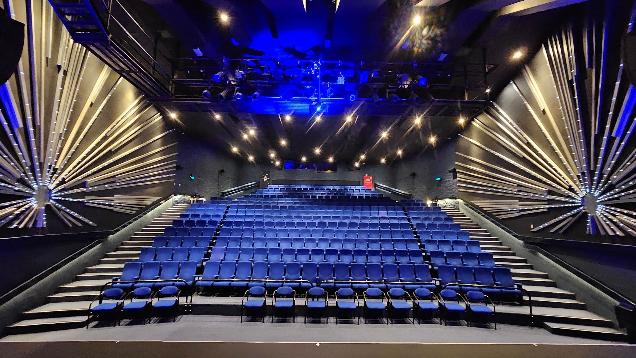 A theatre interior with rows of blue seats arranged in tiers. The walls on both sides feature decorative radial patterns with illuminated lines extending from circular centres. The ceiling is equipped with numerous spotlights and stage lighting, casting a blue hue. The floor is carpeted, and there are aisles on either side of the seating area. A red fire extinguisher is visible on the back wall.