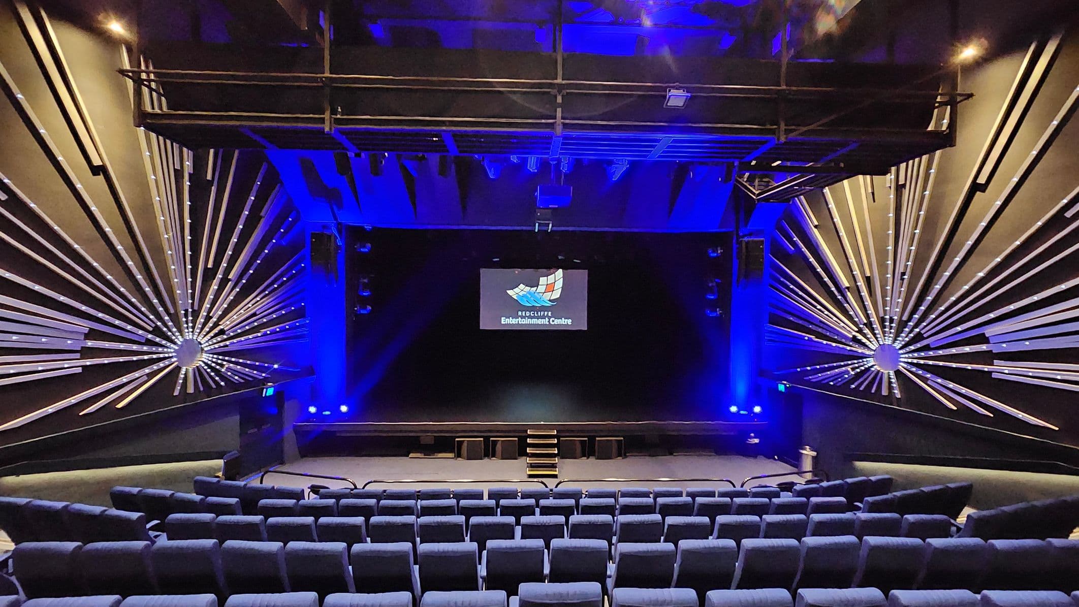 A theatre interior with rows of blue cushioned seats in the foreground. The stage is centred with a small set of steps leading up to it. Above the stage, a screen displays a logo and text. The walls on either side feature decorative radial patterns with illuminated lines extending outward. The ceiling is dark with blue lighting and visible structural elements.