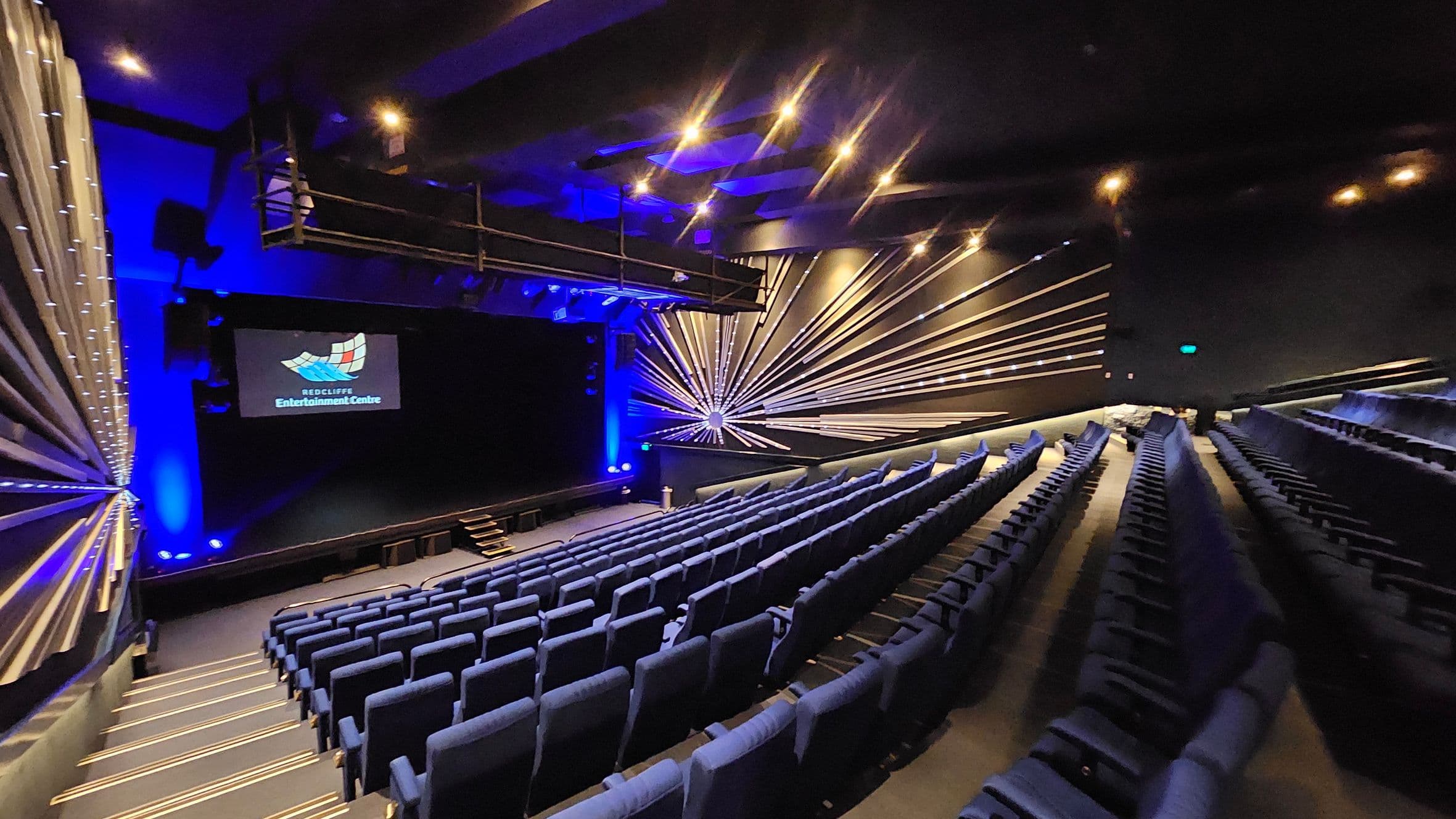 A theatre interior with rows of blue seats arranged in a tiered fashion on the right. The stage is at the front with a screen displaying "Redcliffe Entertainment Centre". The walls feature a decorative design with radiating lines and blue lighting. The ceiling has recessed lighting, and there is a balcony or technical area above the stage. The floor is carpeted with illuminated steps leading down to the stage.