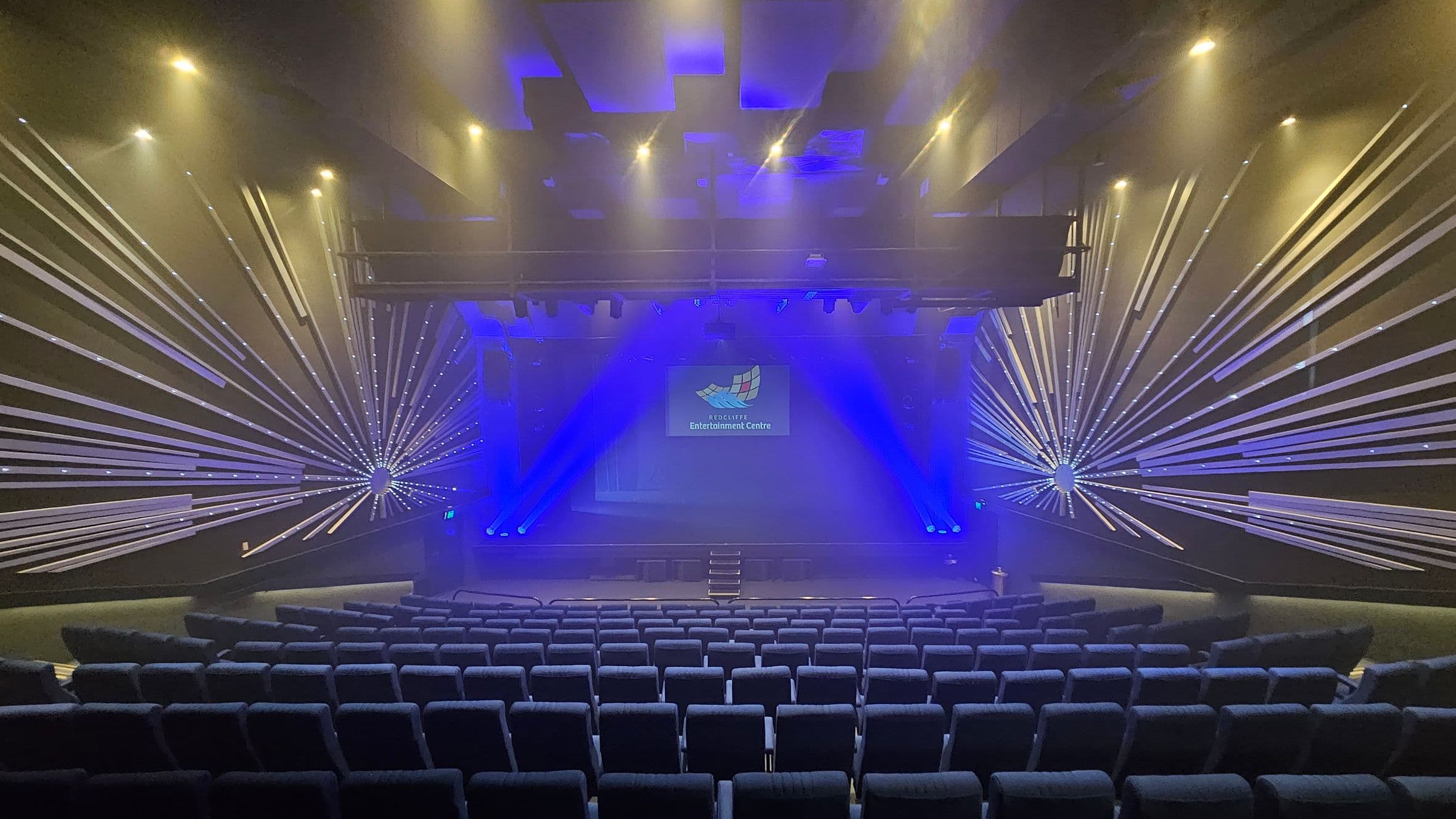 A theatre interior with rows of blue upholstered seats in the foreground. The stage is in the centre, illuminated by blue lights and featuring a screen displaying a logo. The walls on either side have decorative patterns with radiating lines, creating a starburst effect. The ceiling is equipped with spotlights, casting a warm glow. The overall atmosphere is enhanced by a misty effect, likely from stage smoke.