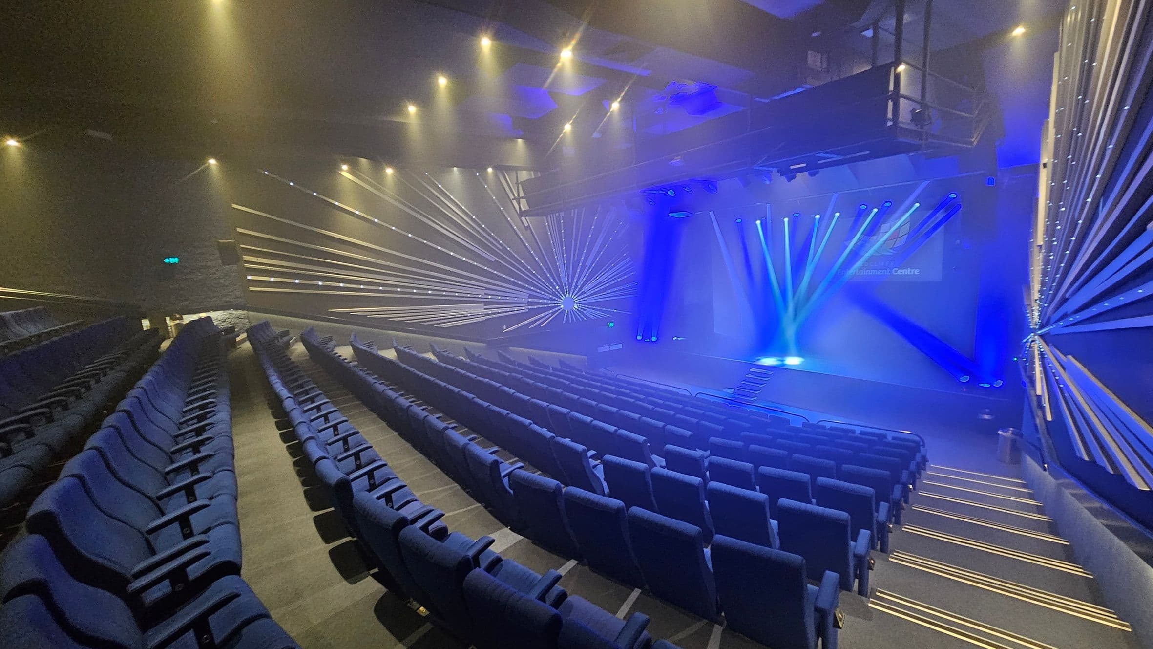 A theatre interior with rows of blue seats arranged in a semi-circular pattern. The stage is illuminated with blue spotlights, and beams of light radiate from the stage towards the audience. The walls have a geometric design with lines converging towards a central point. The ceiling is equipped with multiple spotlights, and the floor is carpeted with steps leading down to the stage.