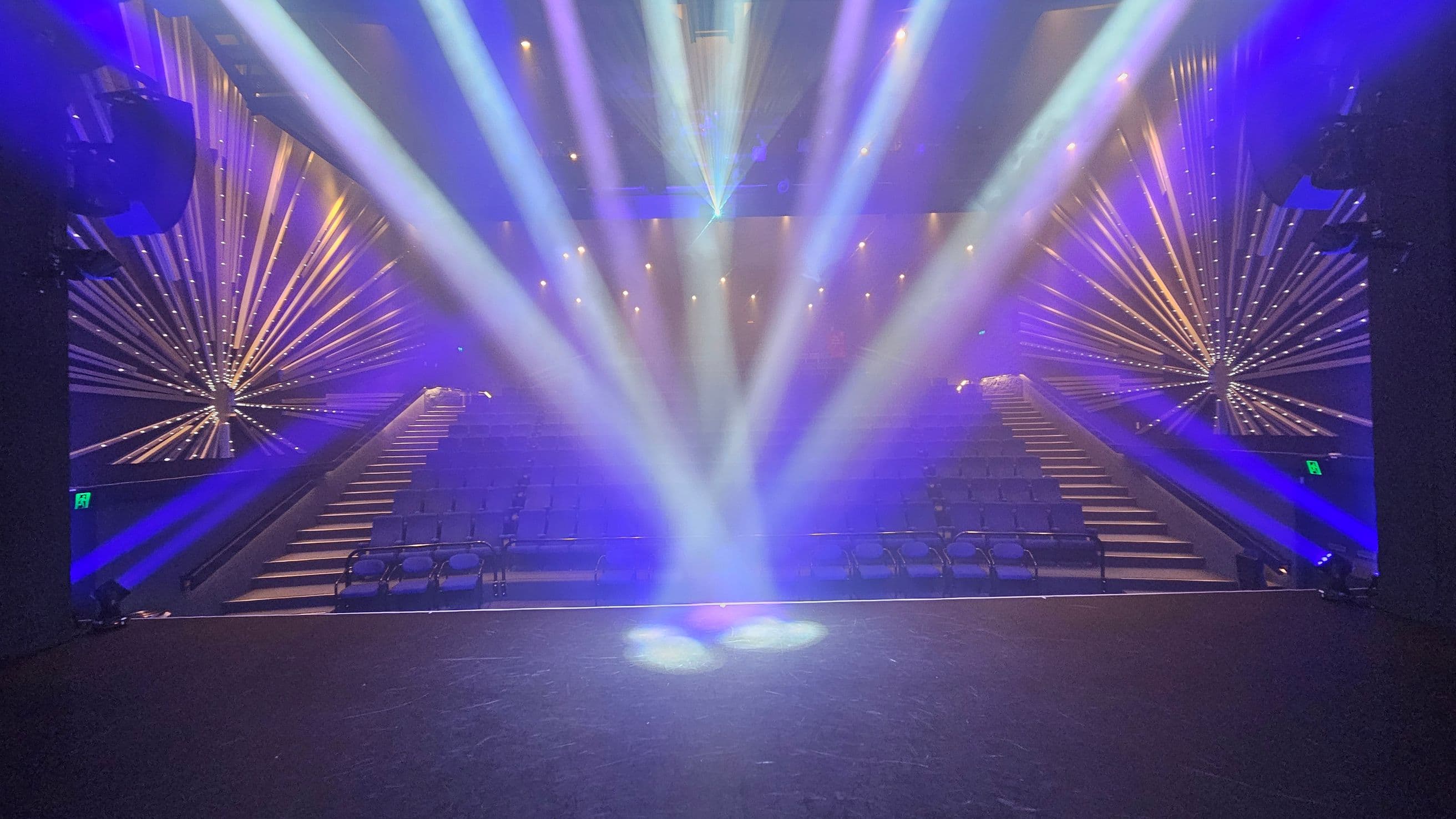 A theatre stage with bright, colourful spotlights shining towards the audience. The seating area is visible, with rows of empty seats. On both sides, there are decorative starburst patterns with illuminated lines extending outward. The ceiling is dark, with additional lights creating a vibrant atmosphere. The floor of the stage is dark and smooth.