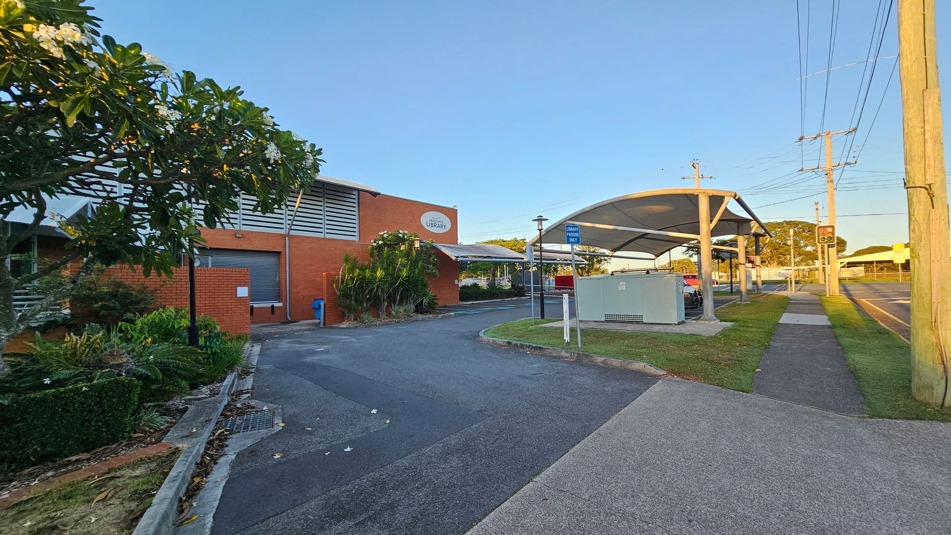 A brick building with a sign indicating it's a library is on the left, surrounded by lush greenery and a tree with white flowers. A paved driveway leads to the building. On the right, there's a covered walkway with a sign for library parking, bordered by a grassy area and a footpath. Utility poles and traffic lights are visible along the road in the background. The scene is well-lit, suggesting it's either early morning or late afternoon.