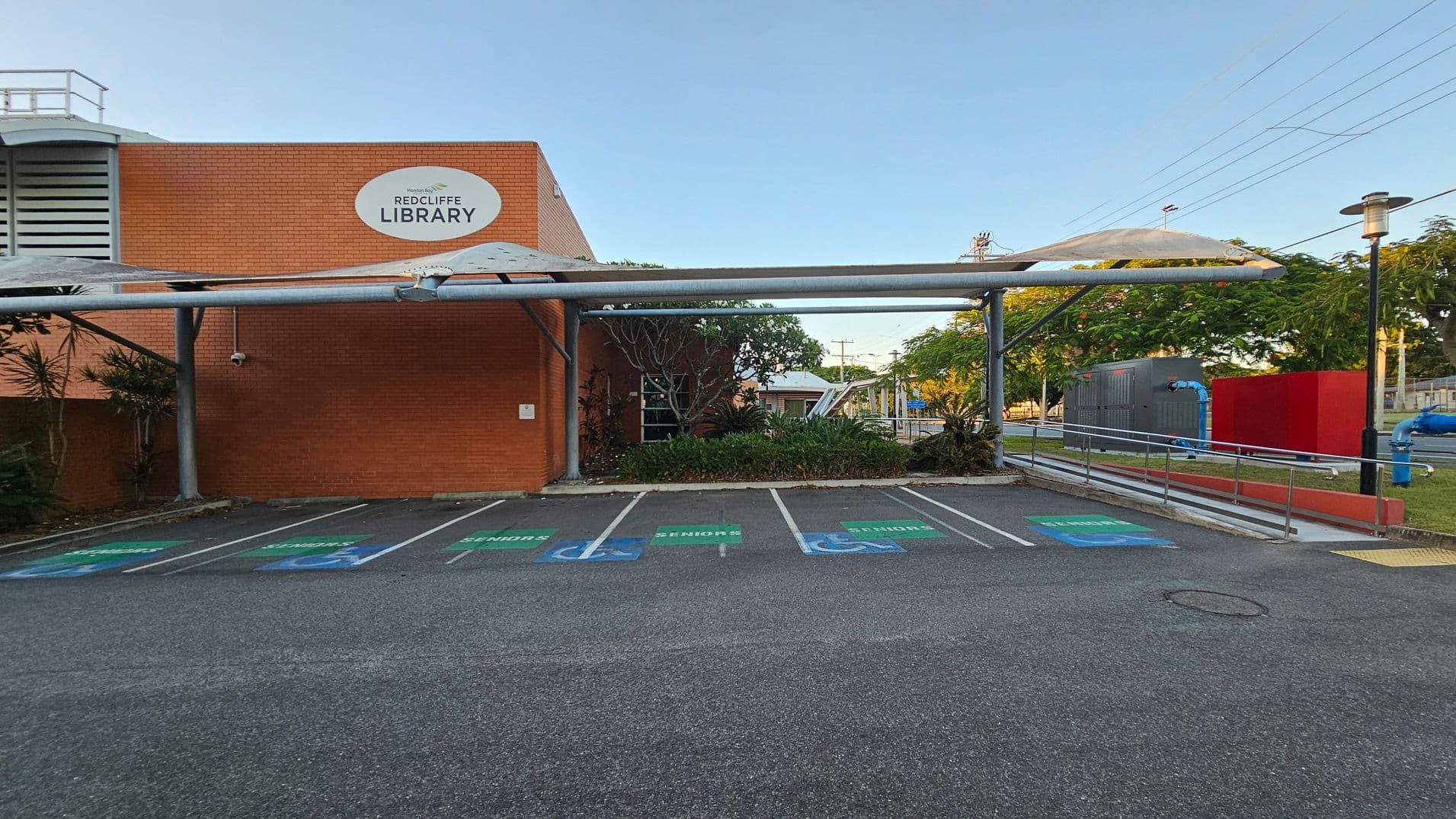 A brick building with a sign reading "Redcliffe Library" on the left. In front, there are several parking spaces marked for seniors and disabled access. A metal canopy extends over the parking area. On the right, there is a ramp with railings leading to the entrance, surrounded by greenery and trees. A red wall and some blue pipes are visible in the background, along with a streetlamp. The ground is asphalt, and the area is well-maintained.