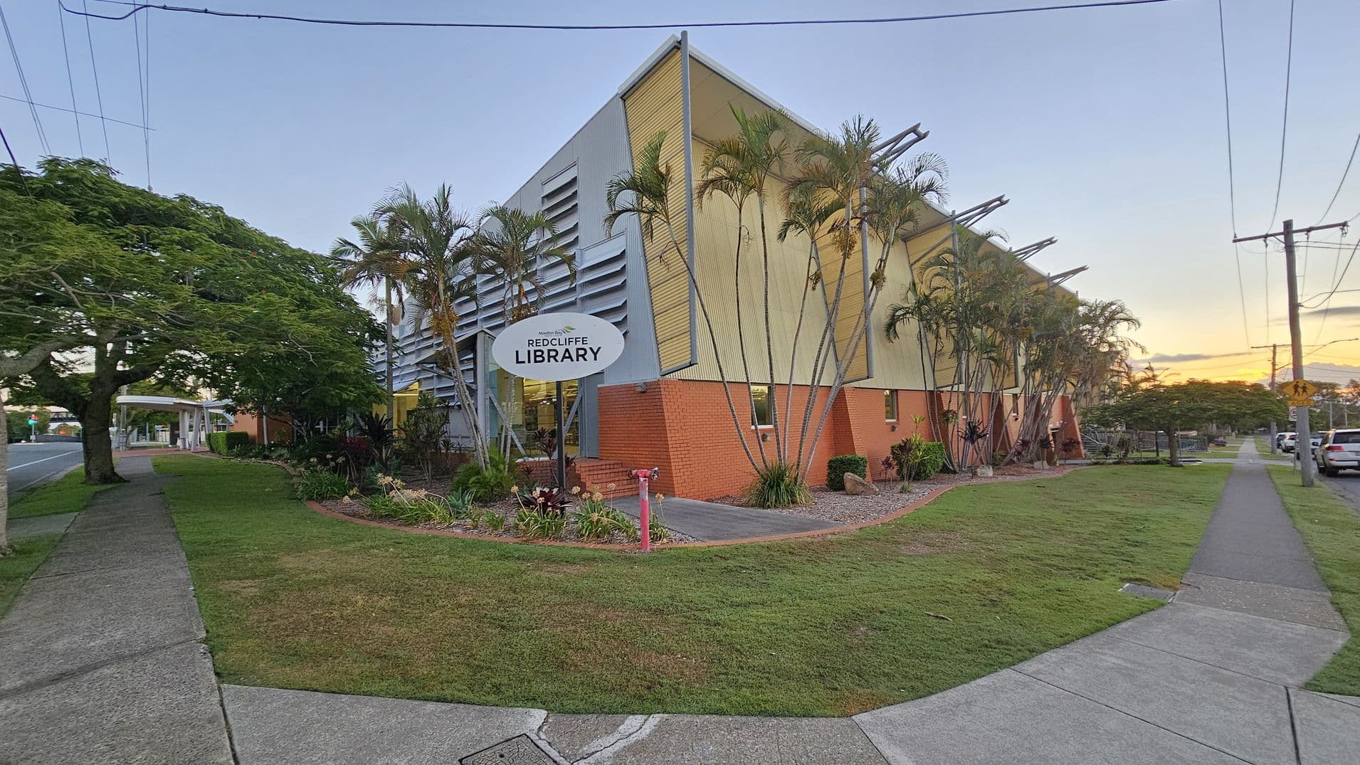 A modern building with a slanted roof and a mix of brick and metal siding, identified as "Redcliffe Library" on a sign in front. Tall palm trees line the side of the building. The foreground features a grassy area with a concrete footpath leading to the entrance. On the left, a large tree provides shade over the footpath, which runs parallel to a road. On the right, a row of parked cars is visible along the street. Power lines stretch across the top of the image.