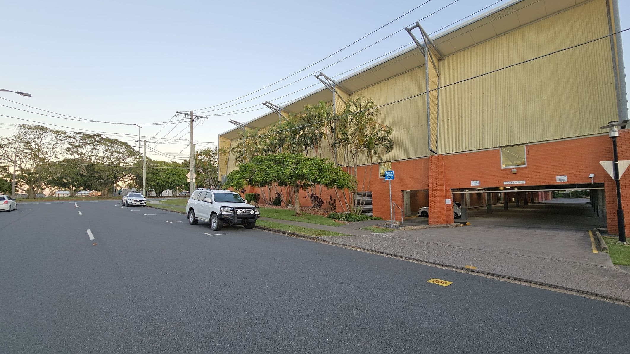 A street scene with a large building on the right, featuring a red brick lower section and a beige upper section. Several palm trees and shrubs are in front of the building. A white SUV is parked on the street, with other cars visible in the distance. The road is wide with marked lanes, and power lines run overhead. The background includes more trees and a clear sky. A covered parking area is visible under the building.