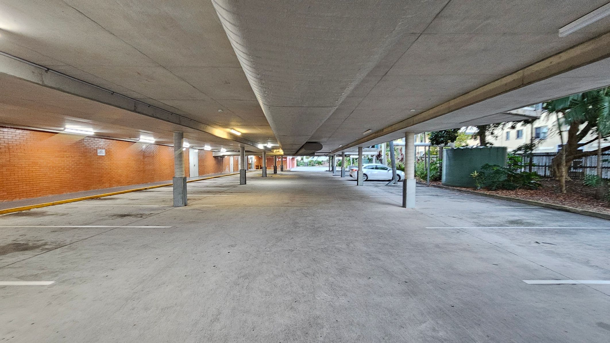 A covered parking area with a concrete floor and ceiling. On the left, there is a long brick wall with several lights illuminating the space. The right side features a row of parking spaces, with a white car parked near some greenery and a large cylindrical water tank. Several support columns run down the centre, and the area is open at the far end, leading to an outdoor space with trees and buildings visible in the background.