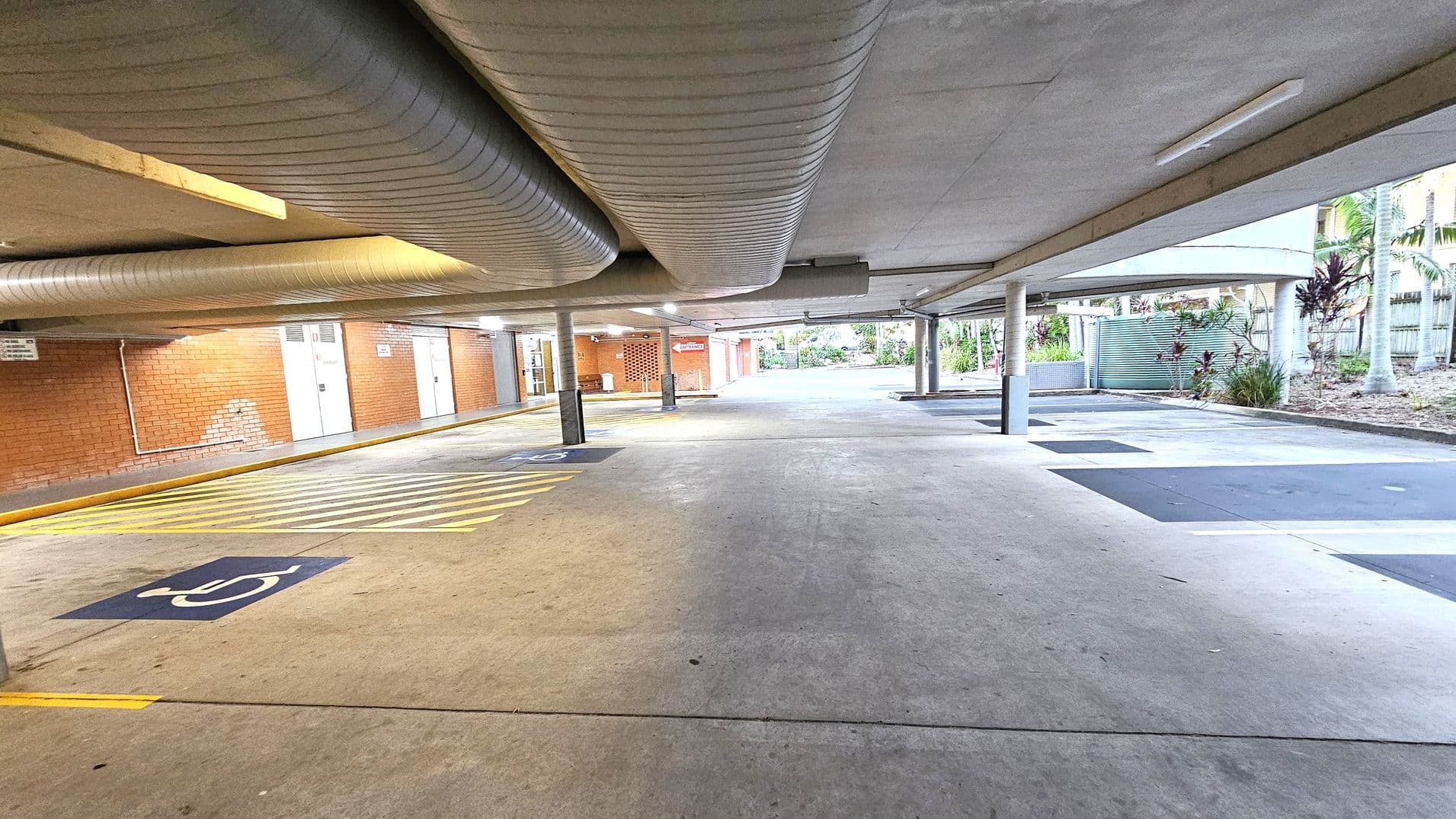 A covered parking area with a concrete floor and ceiling. On the left, there are brick walls with several doors and a sign. Yellow lines and a disabled parking symbol are painted on the ground. Large ventilation ducts run along the ceiling. On the right, there are open spaces with some greenery and palm trees visible outside. Several support columns are evenly spaced throughout the area.