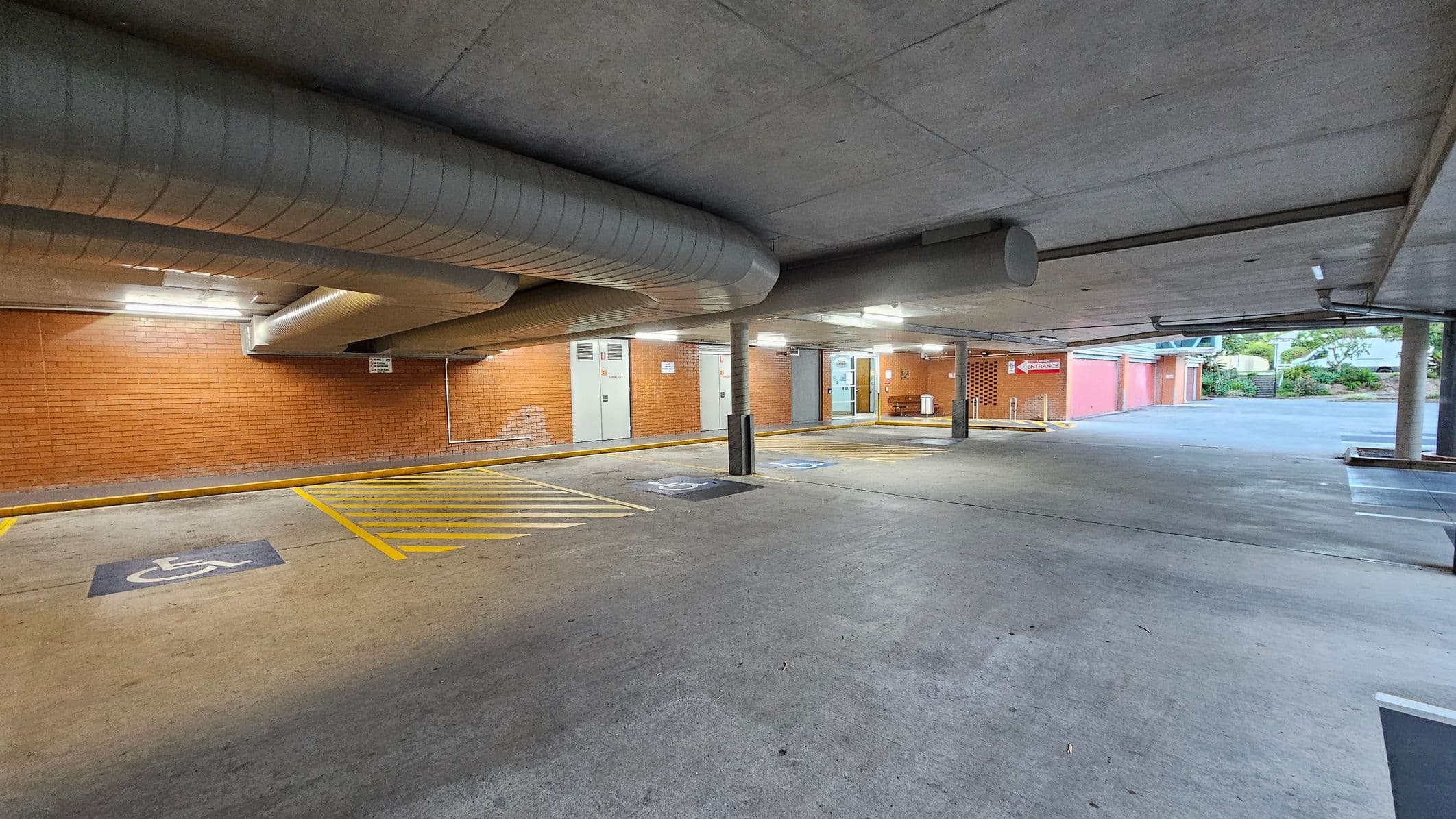 Underground car park with a concrete floor and ceiling. On the left, there are designated disabled parking spaces marked with yellow lines and symbols. The brick wall in the background has several doors and a sign indicating an entrance. Large ventilation ducts run along the ceiling. On the right, the car park opens to an outdoor area with greenery and a staircase leading upwards. The lighting is provided by overhead fluorescent lights.