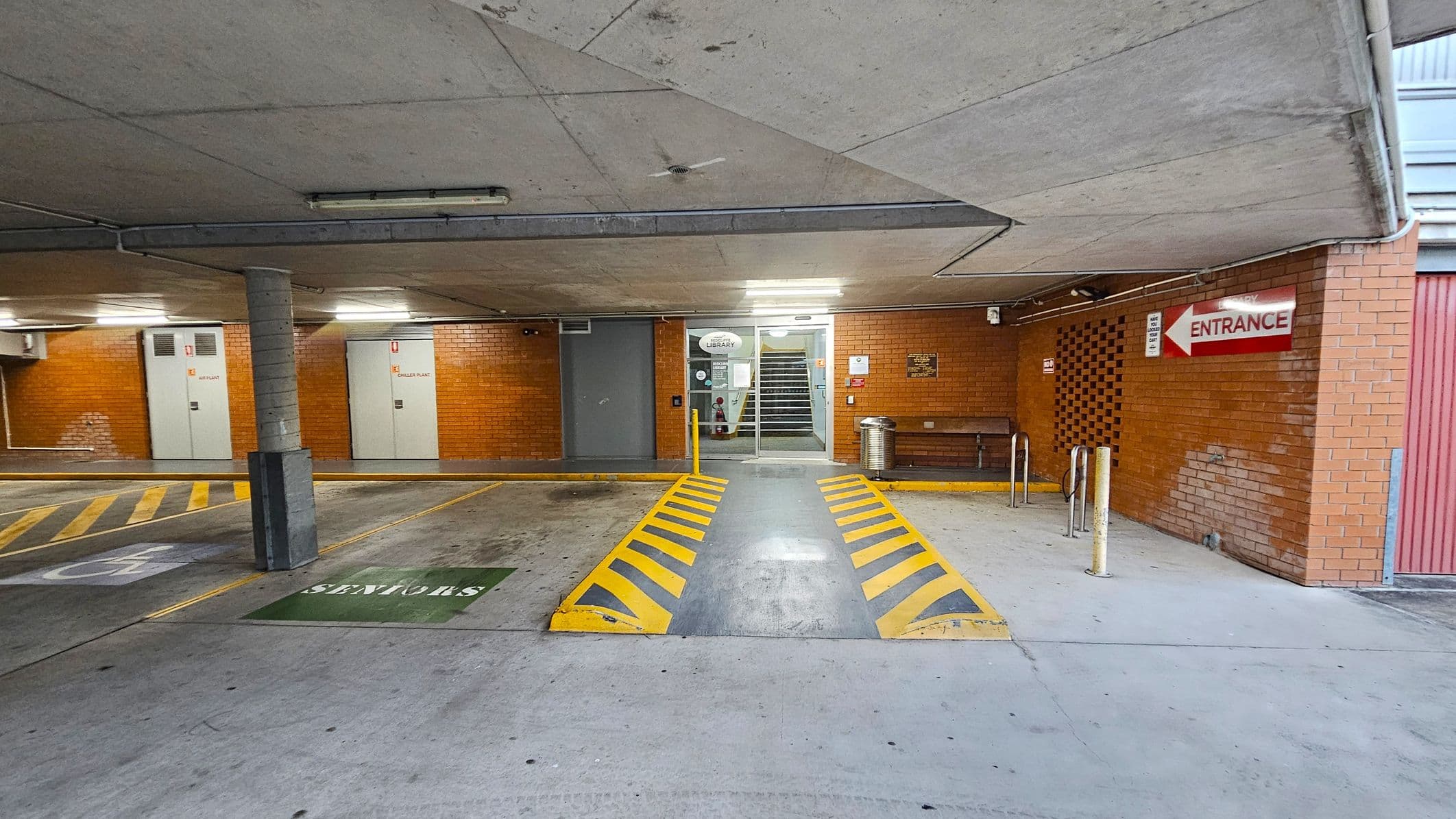 A concrete parking area with a ramp in the centre marked by yellow and black stripes. On the left, there are parking spaces with a "Seniors" sign and a pillar. The background features brick walls with doors labelled "Diesel Plant" and "Library" near a staircase. On the right, a red sign indicates "Entrance" above a bench and a metal bin. The ceiling is concrete with fluorescent lights.
