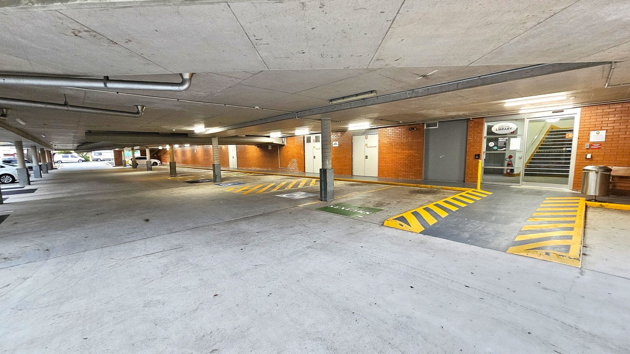 A covered car park with a concrete floor and ceiling. On the right, there is a brick wall with a glass door leading to a staircase, marked with a "Library" sign. Yellow and black striped markings are on the ground near the entrance. Several parking spaces are visible, with a few cars parked on the left. The area is lit with fluorescent lights, and there are metal pipes running along the ceiling. A metal bin and a bench are near the entrance.