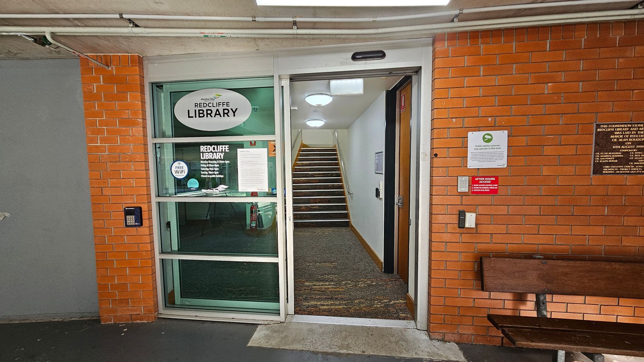 A brick wall with a glass door on the left, displaying "Redcliffe Library" signage and information about free Wi-Fi. The door is open, revealing a carpeted staircase leading upwards. On the right, there are plaques and signs on the wall, including one about after-hours access. A wooden bench is positioned against the wall. The ceiling has exposed pipes and lights, and the floor is concrete.