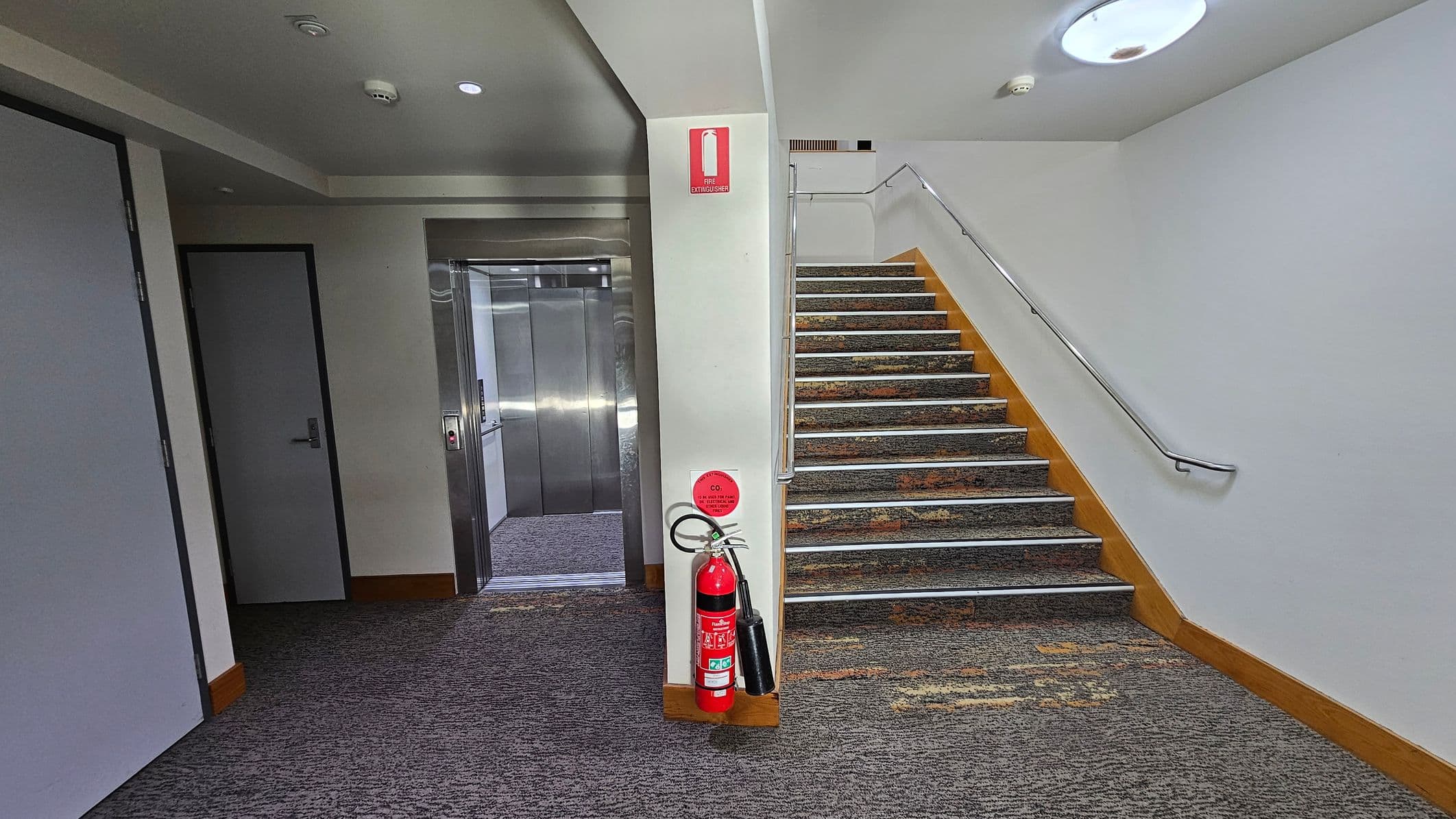 A hallway with a grey carpeted floor. On the left, there is a closed door and an open lift with metallic doors. In the centre, a fire extinguisher is mounted on a white wall. To the right, a staircase with a patterned carpet and a metal handrail ascends. The ceiling is white with recessed lighting and a smoke detector.