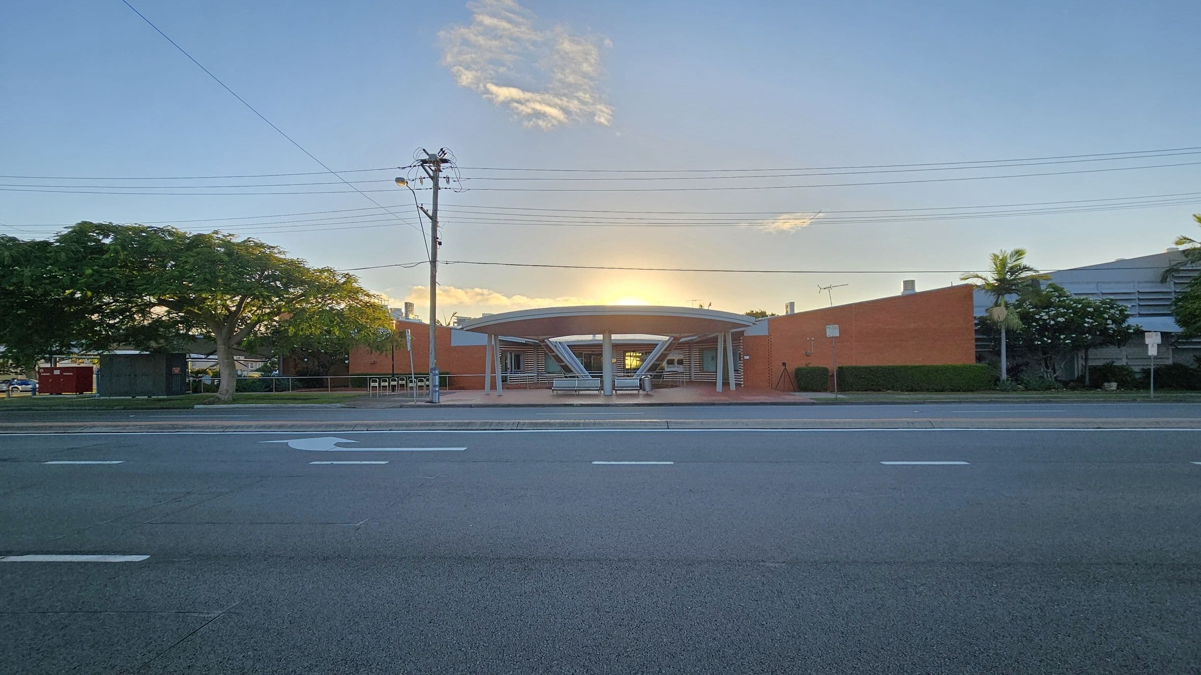 A wide road in the foreground with white lane markings. In the middle, a building with a circular entrance and a flat roof, flanked by red brick walls. A large tree with a thick trunk and green foliage is on the left, while smaller trees and shrubs are on the right. Overhead, power lines stretch across the scene. The sun is setting behind the building, casting a warm glow.