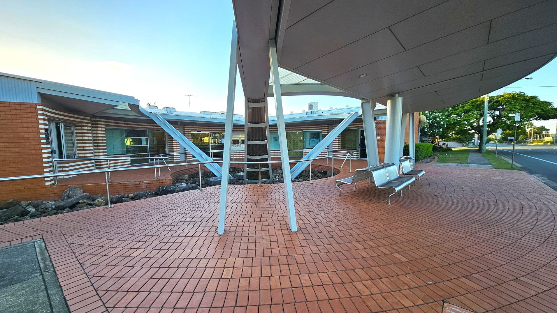 A modern building with a curved design featuring red brick walls and white horizontal stripes. The foreground has a tiled area with metal benches. The structure is supported by angled metal beams. A glass window displays the word "LIBRARY" inside. To the right, a road runs alongside a grassy verge with trees and a lamppost. The ceiling is smooth and white, with recessed lighting.