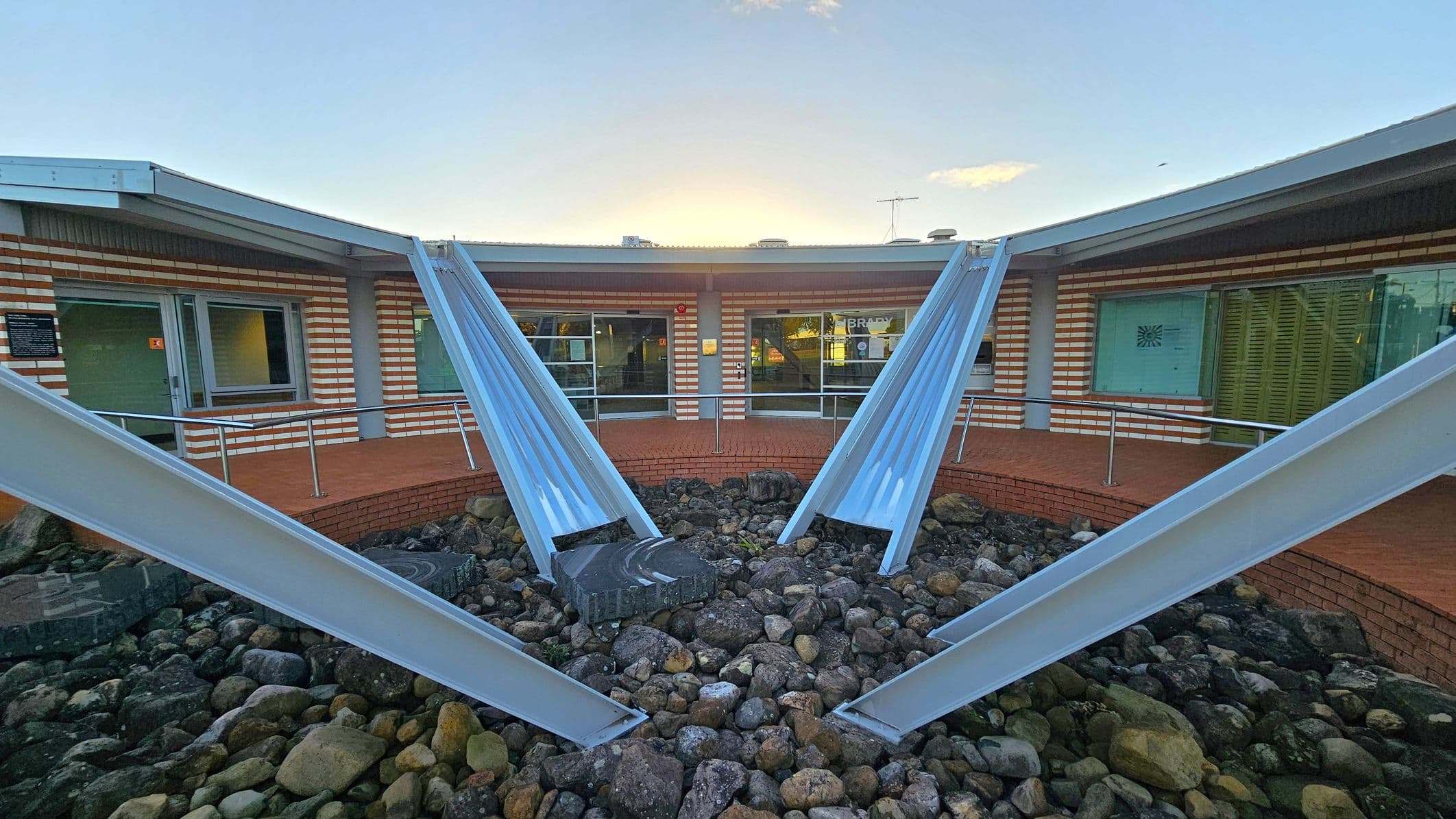 A courtyard with a circular arrangement of metal beams converging towards the centre, surrounded by a bed of rocks. The background features a building with a brick facade and large windows. The building has a flat roof with a metal edge. The ground is covered with stones, and there is a small raised brick platform near the building. The sky is visible above the structure.