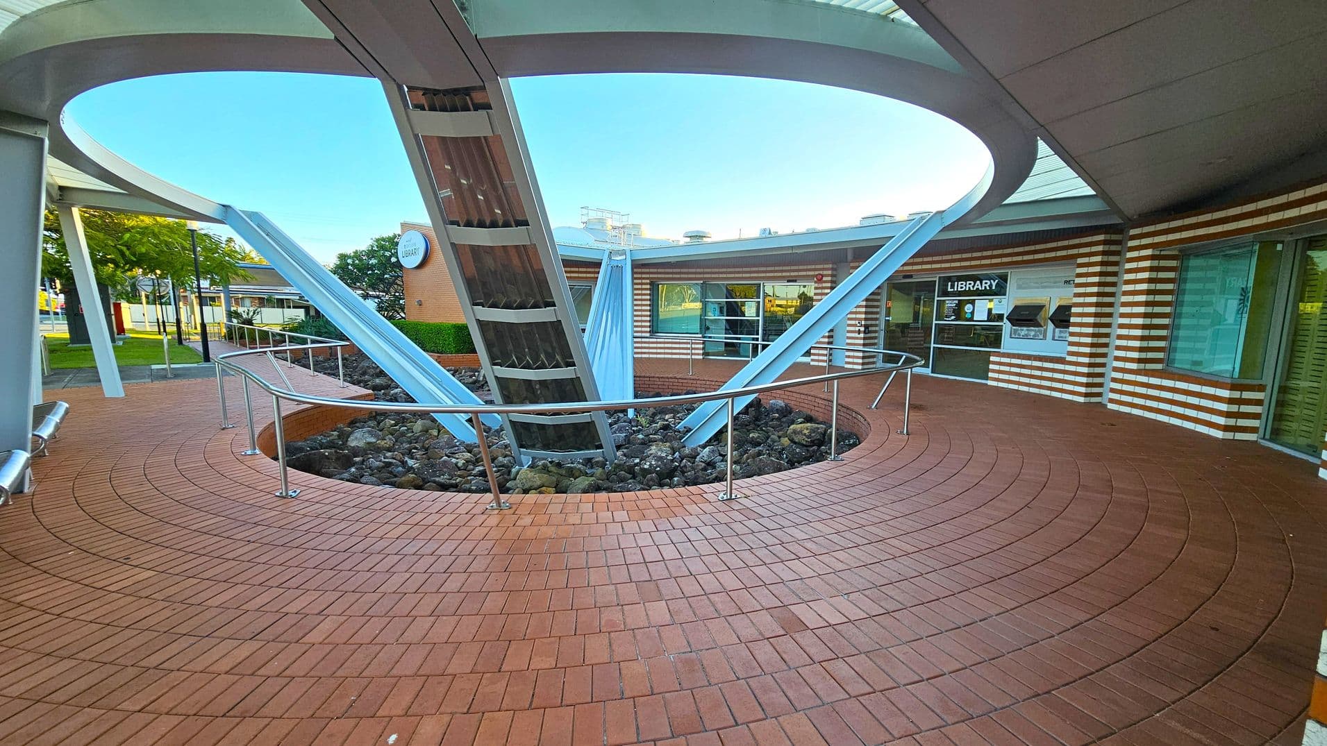 Circular courtyard with a brick floor and a central metal structure featuring angled beams. A railing surrounds a rocky area at the base of the structure. The background shows a building with striped brickwork and large windows, one marked "Library". Trees and a street are visible to the left. The ceiling is a curved metal canopy with an open circular section above the structure.