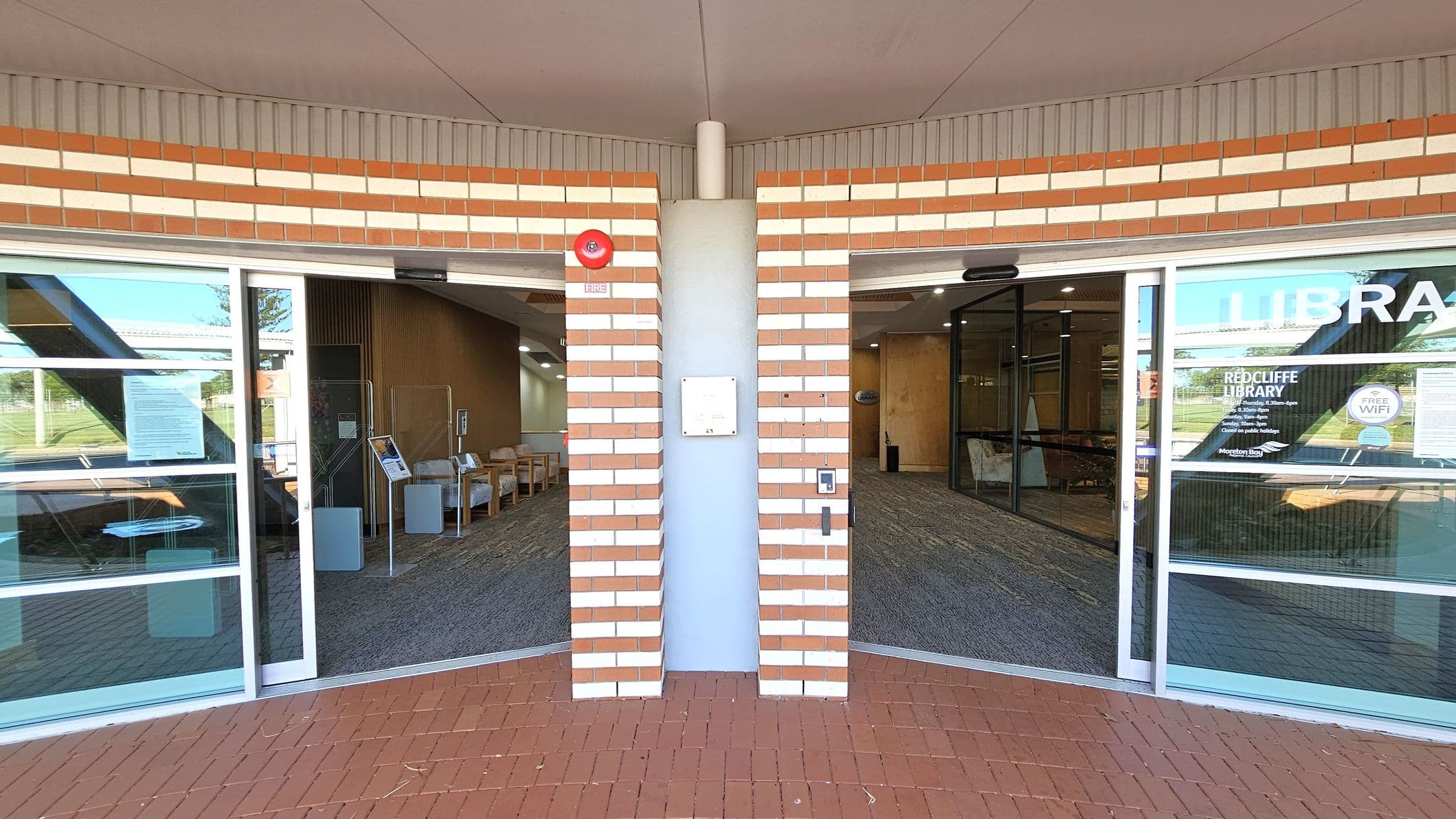 A brick wall with alternating red and white bricks is in the centre, featuring a fire alarm and a small sign. On either side, there are glass doors leading into a library. The left side shows a seating area with chairs and a notice board. The right side has signage for "Redcliffe Library" and "Free WiFi". The floor is covered with red tiles outside and a patterned carpet inside. The ceiling is plain with a central seam.