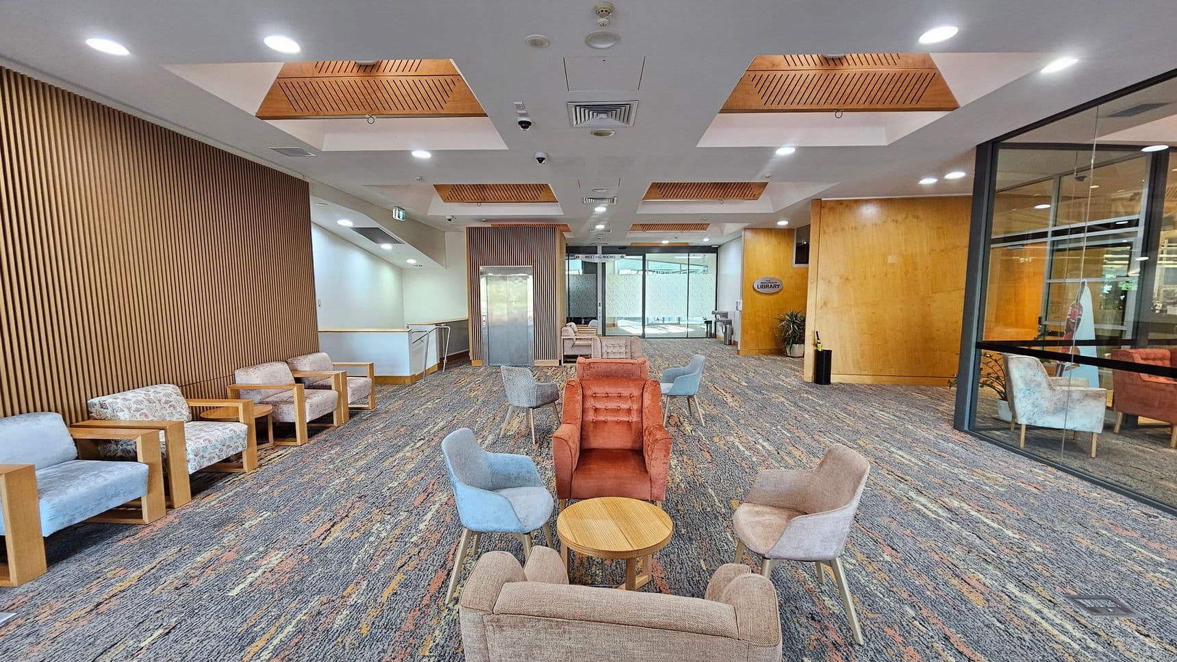 A spacious lobby area with a patterned carpet floor. On the left, there is a row of cushioned chairs with wooden frames against a wall with vertical wooden slats. In the centre, a cluster of colourful armchairs surrounds small round tables. The ceiling features recessed lighting and wooden panels. On the right, a glass wall reveals more seating and a sign indicating a library. An elevator is visible in the background, flanked by more seating.
